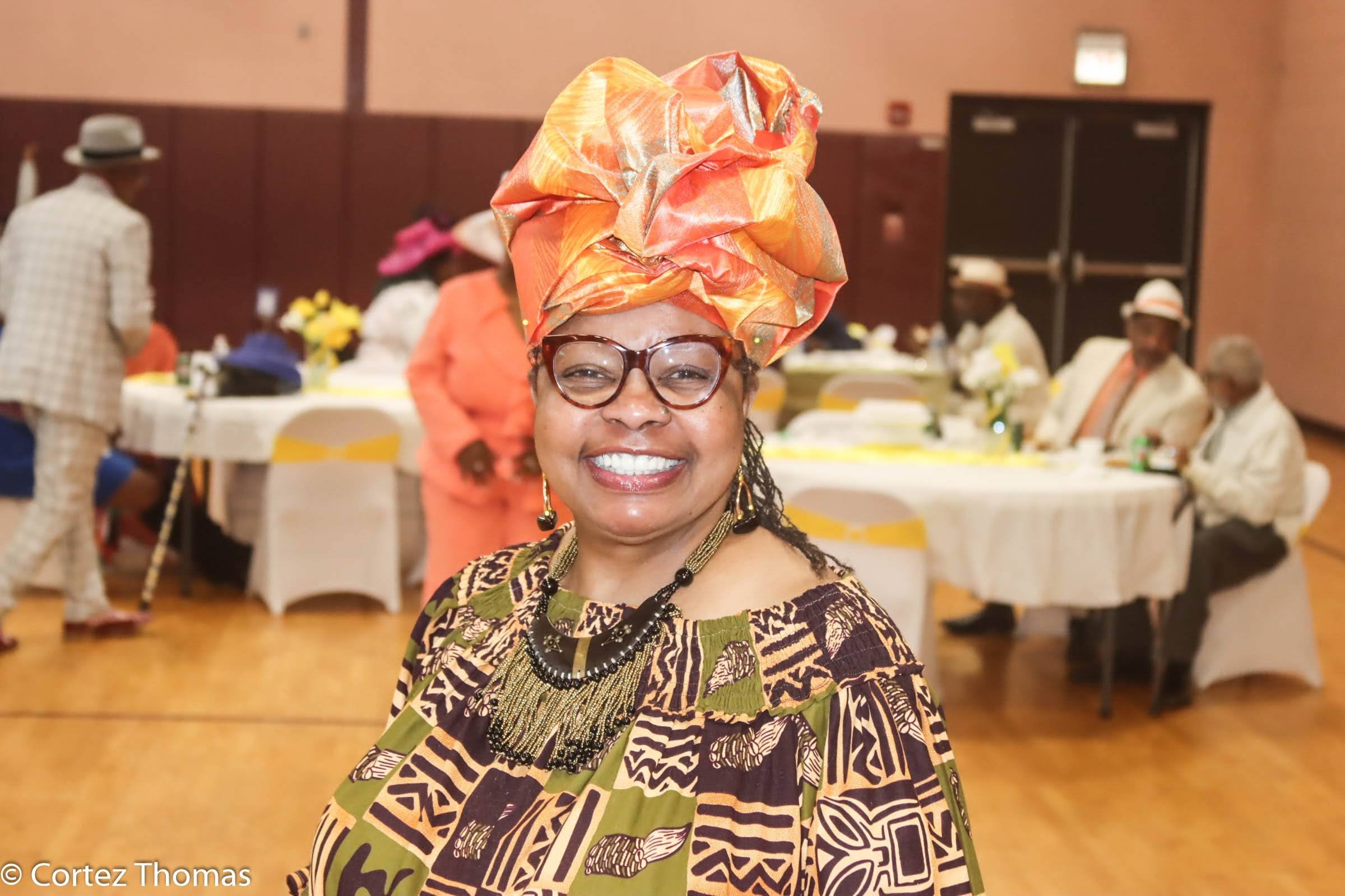 A smiling woman wearing glasses, an orange headwrap, and a colorful African print dress standing in a decorated event hall with guests seated at tables in the background.
