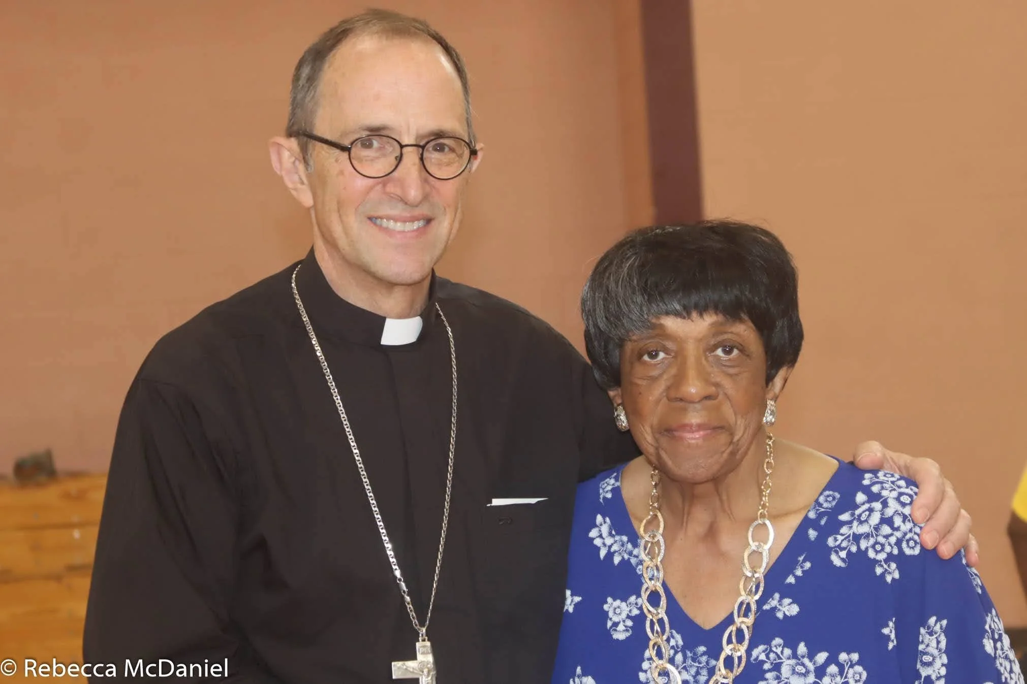 A man dressed as a priest in black with glasses and a cross necklace standing next to an elderly woman in a blue floral top with a gold chain necklace, both smiling and posing for the camera.