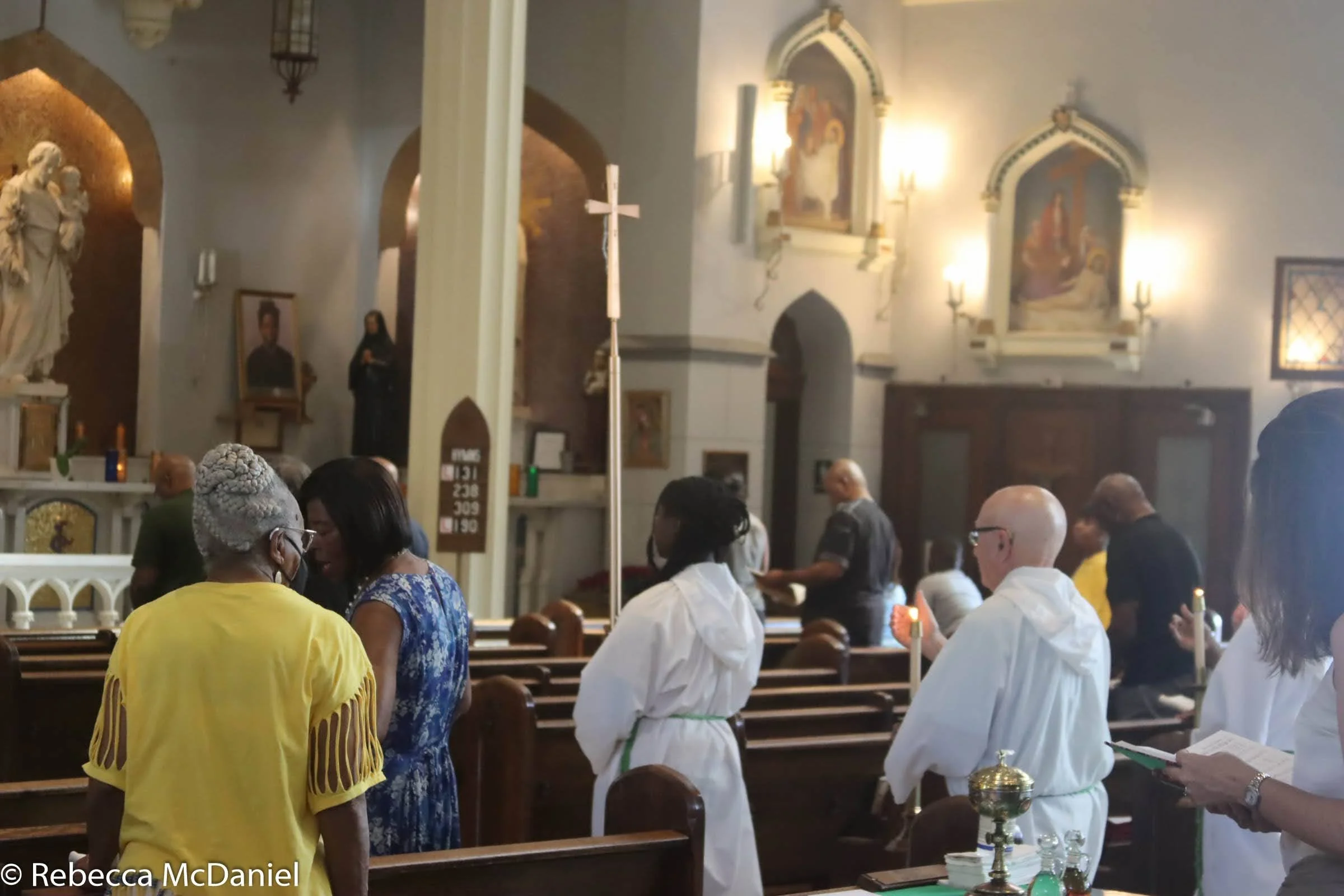 People inside a church, some are seated, and some are standing, with religious statues and paintings on the walls and a tall lit candle in the foreground.
