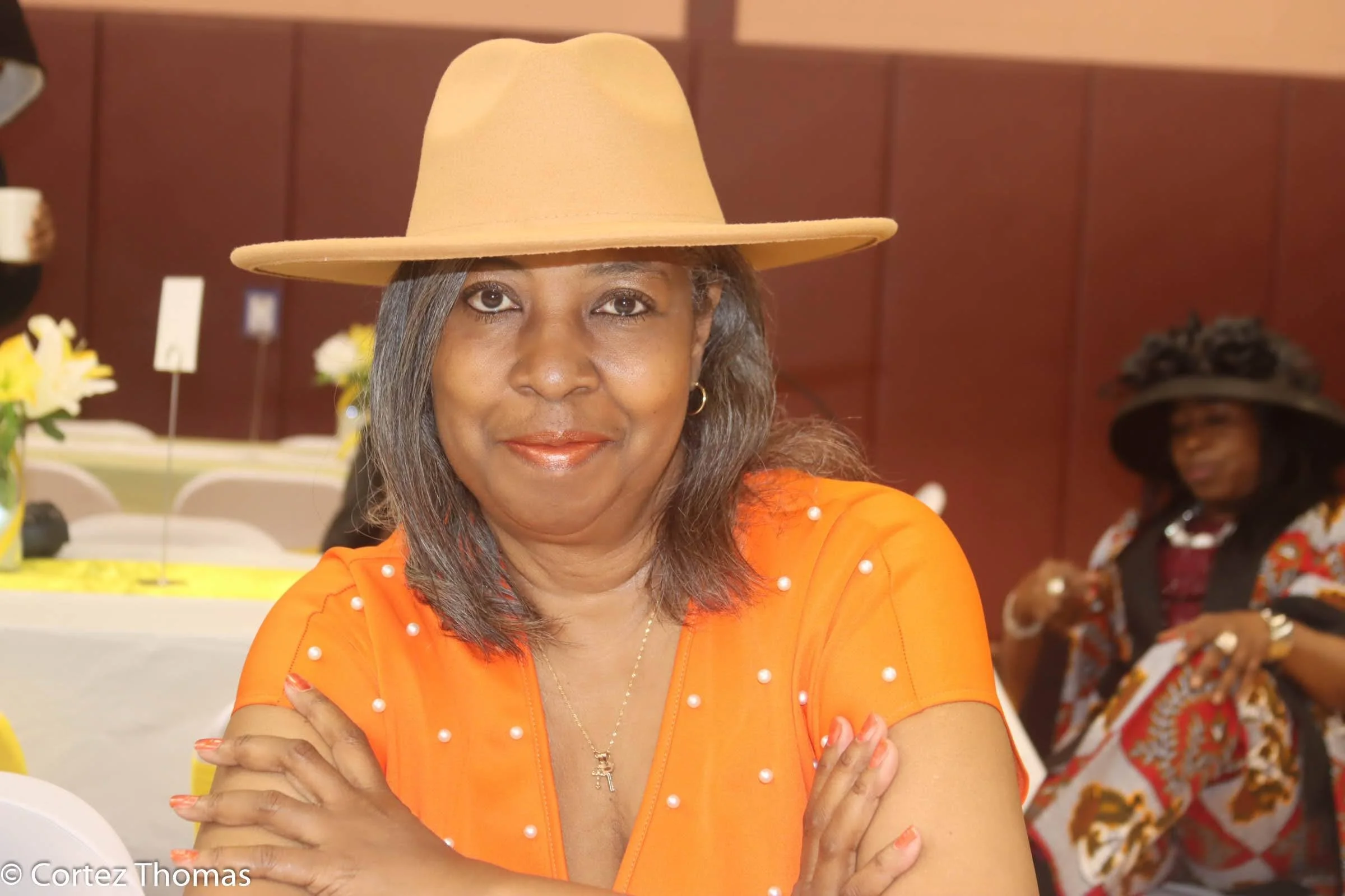 A woman wearing an orange dress with pearl accents, a beige wide-brimmed hat, and a gold necklace, sitting at a table with her arms crossed, smiling at the camera. In the background, another woman wearing a large hat and a colorful patterned dress is