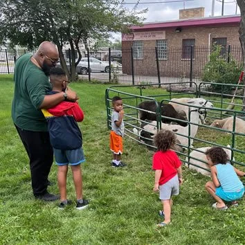 A man with four children at a petting zoo, one child is petting a goat through a metal fence, others are watching or interacting nearby, with trees and a brick building in the background.