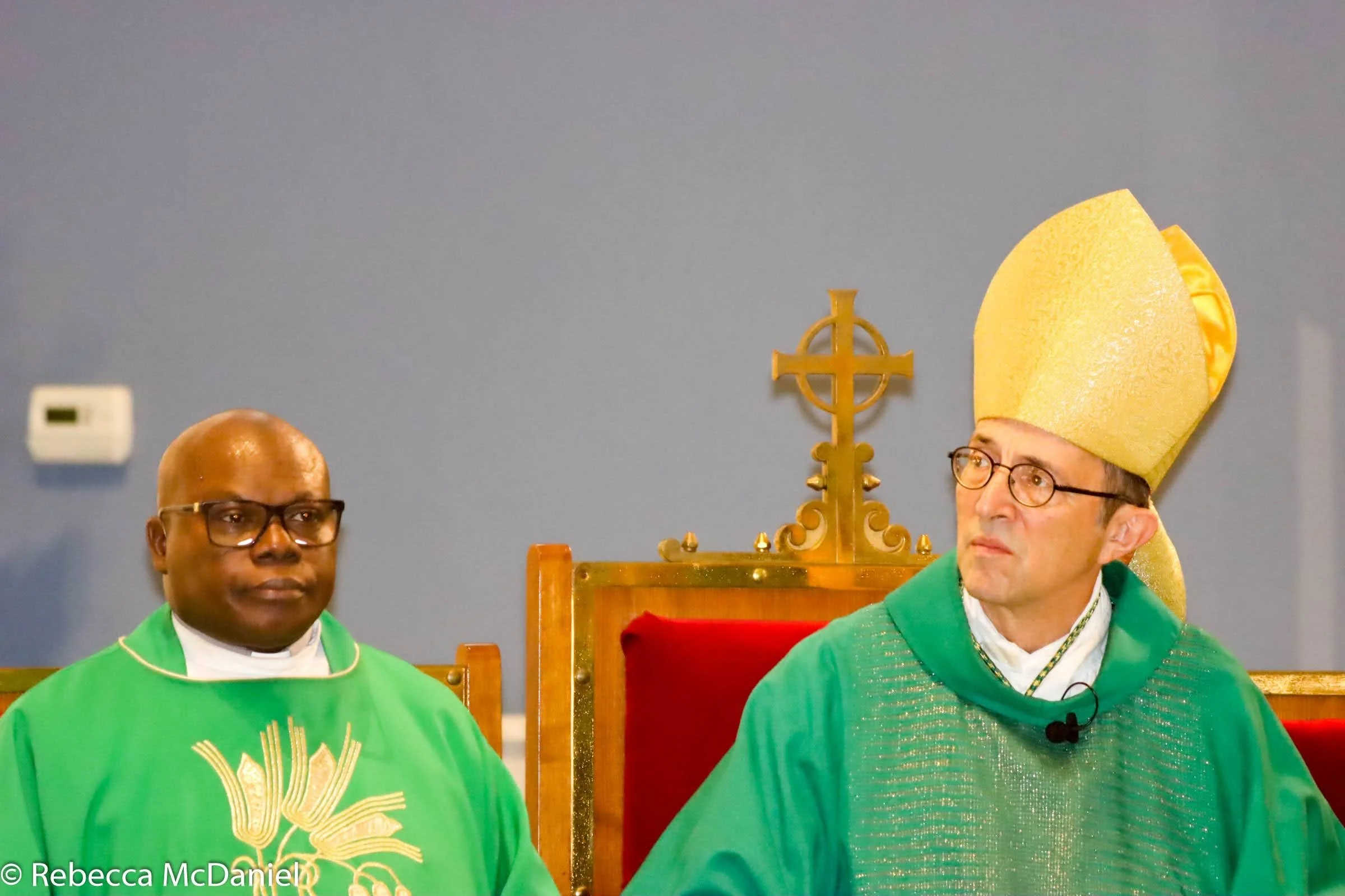 Two clergy members sitting in church, one wearing green vestments and the other wearing a golden mitre and green vestments.