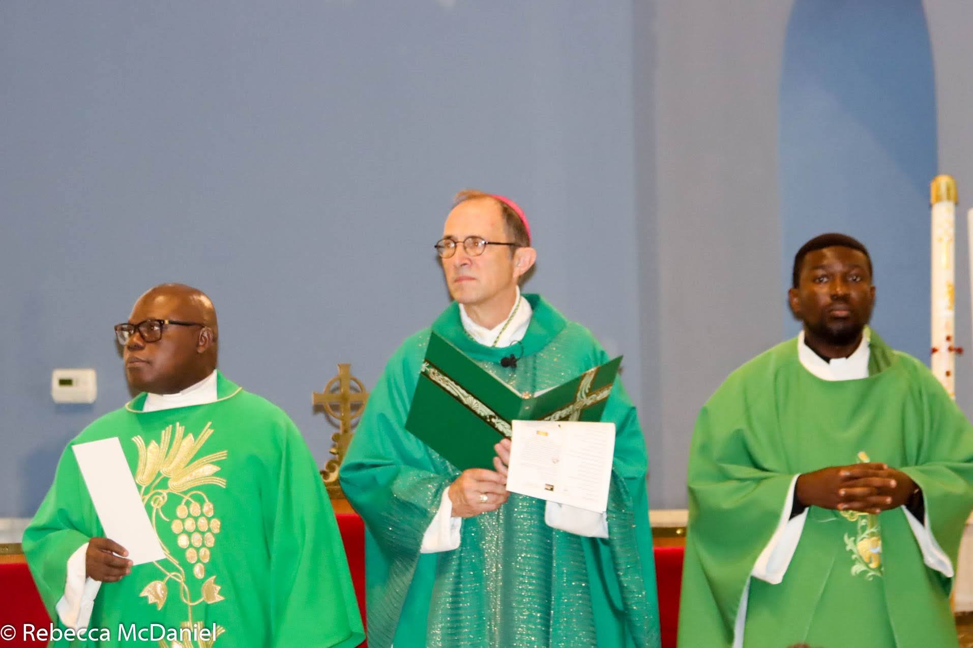 Three clergymen dressed in green liturgical vestments standing in a church, holding religious items during a service.