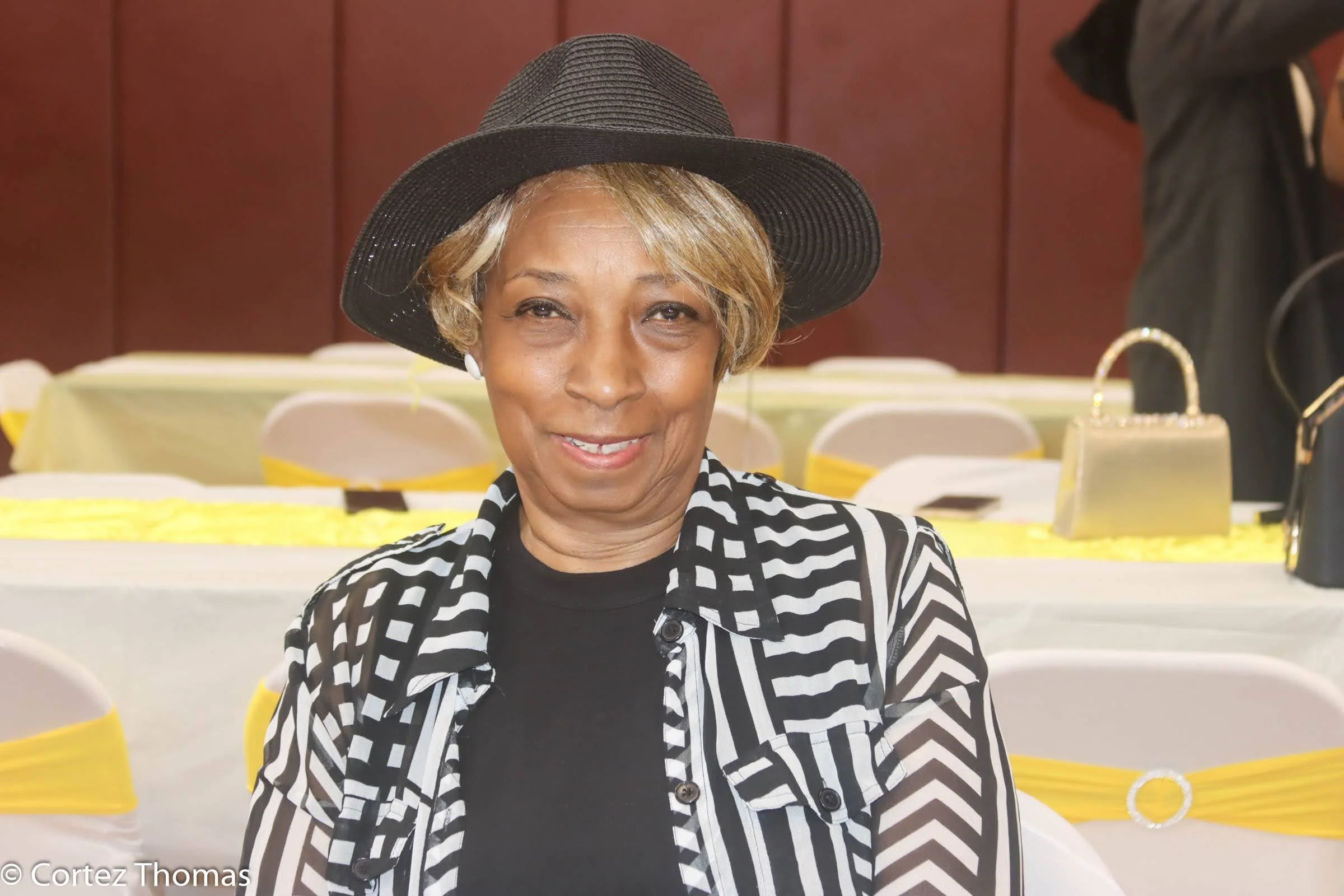 A woman with light brown skin, short blonde hair, wearing a black hat, black and white striped blazer, and black top, smiling at a formal event with decorated chairs in the background.