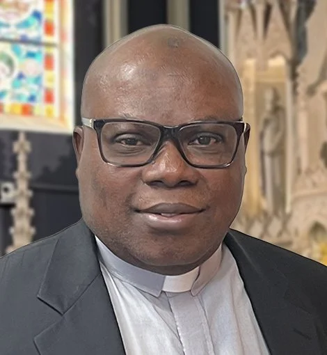 A man wearing a clerical collar, black suit, and glasses, featuring a close-cropped haircut, standing indoors with stained glass and religious decor in the background.