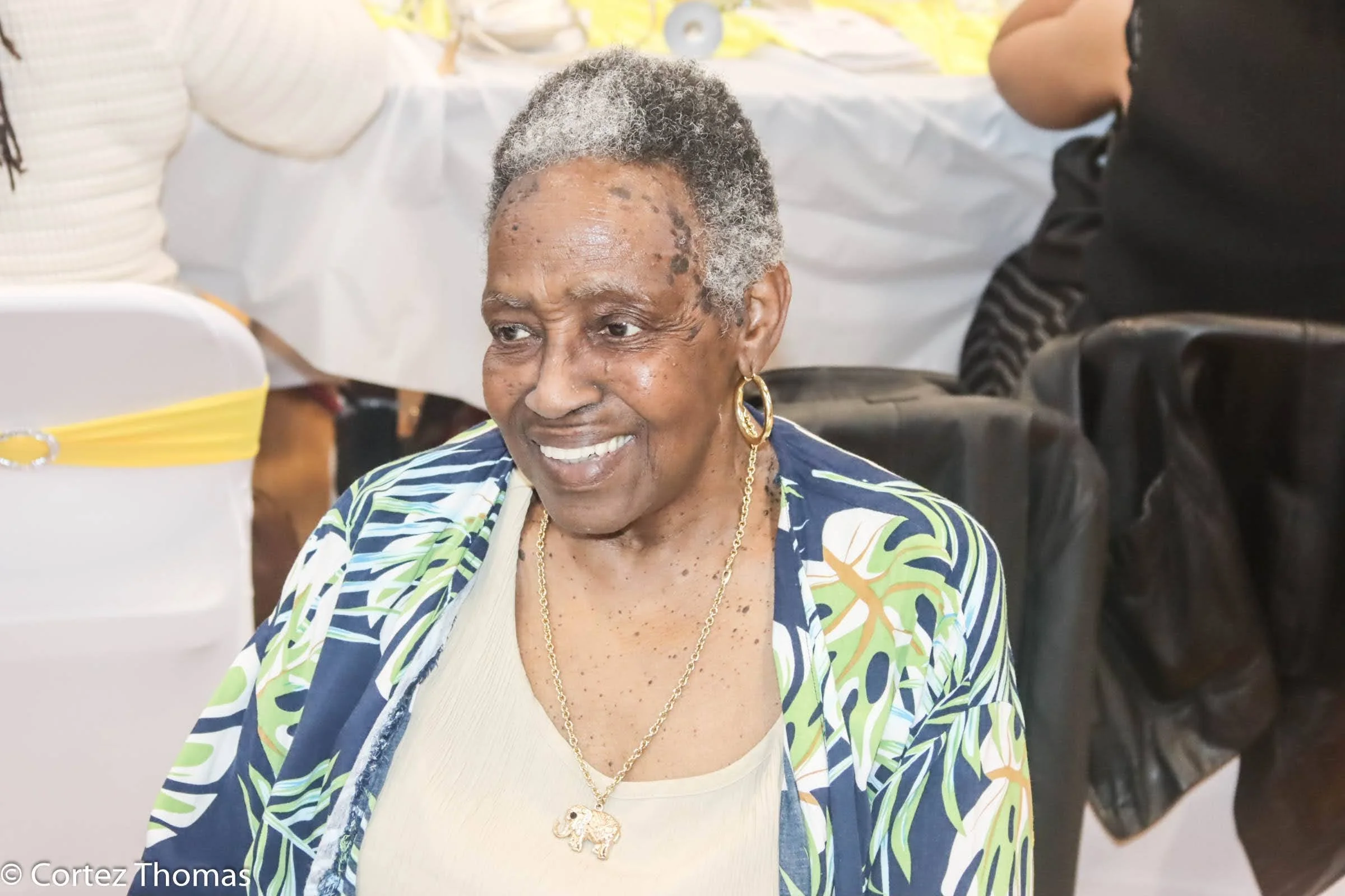 Older woman with short gray hair smiling, wearing a beige top, blue, green, and white patterned jacket, large gold hoop earrings, and a gold necklace with an elephant pendant, sitting at a decorated event.