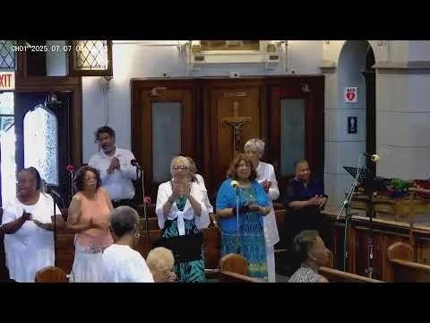 People standing and singing in a church with a priest or minister at a microphone during a service.