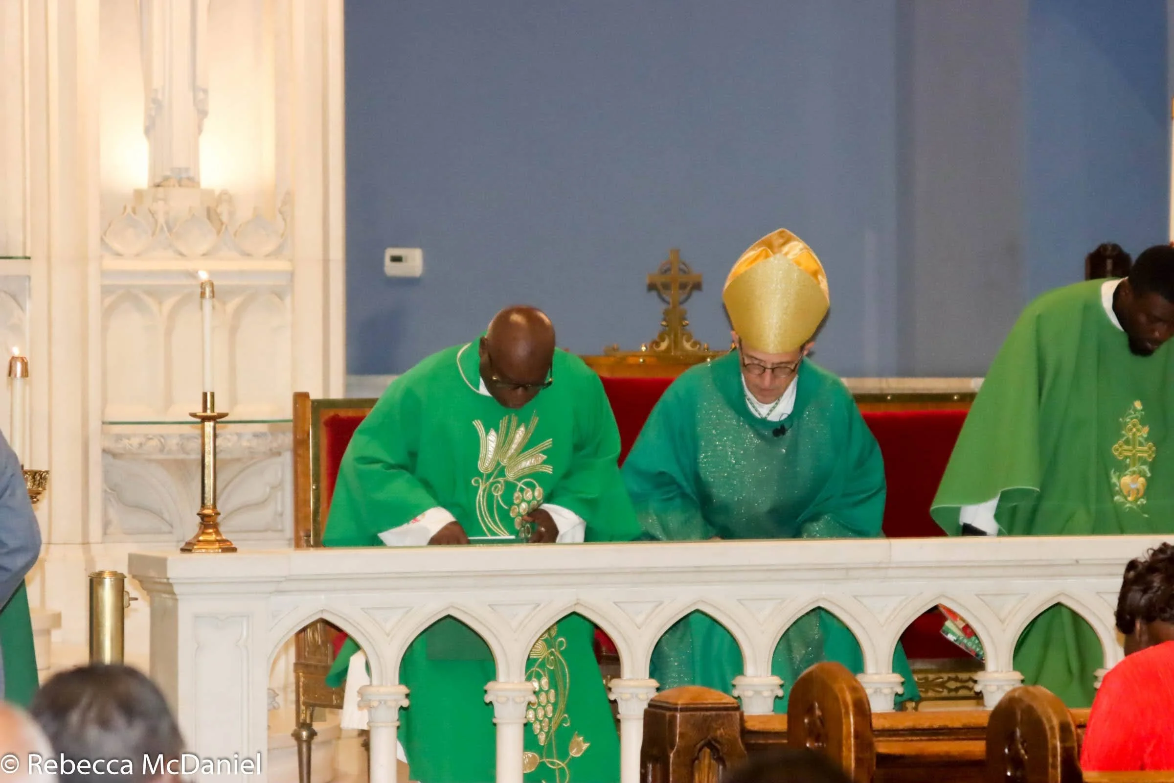 Three clergy members in green robes kneeling at an altar during a religious service inside a church, with one wearing a gold mitre and glasses