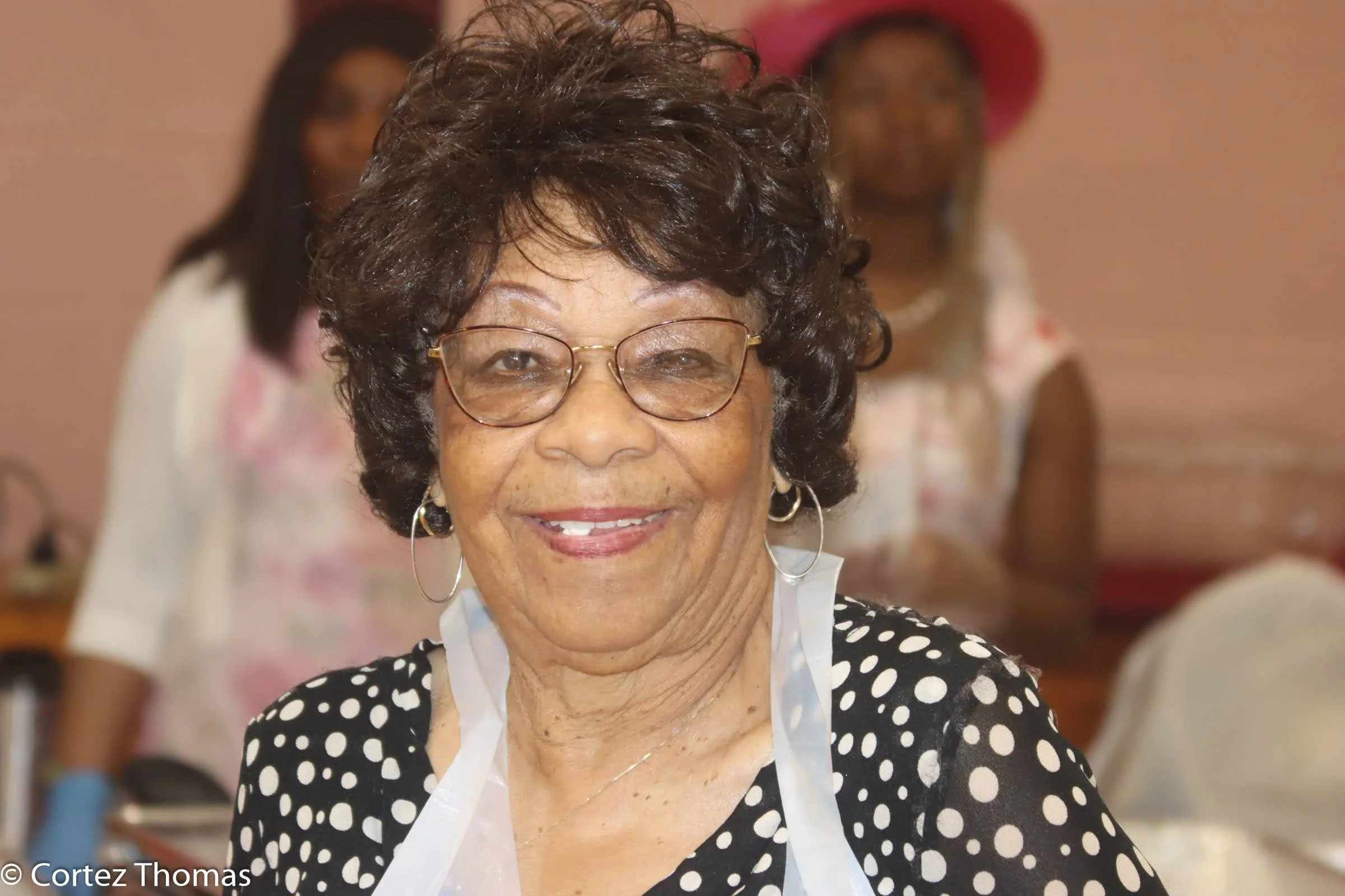 Close-up of an elderly woman with short, curly dark hair, glasses, and hoop earrings, smiling at the camera. She is wearing a black and white polka dot blouse with a white collar. Blurred women are seen in the background.