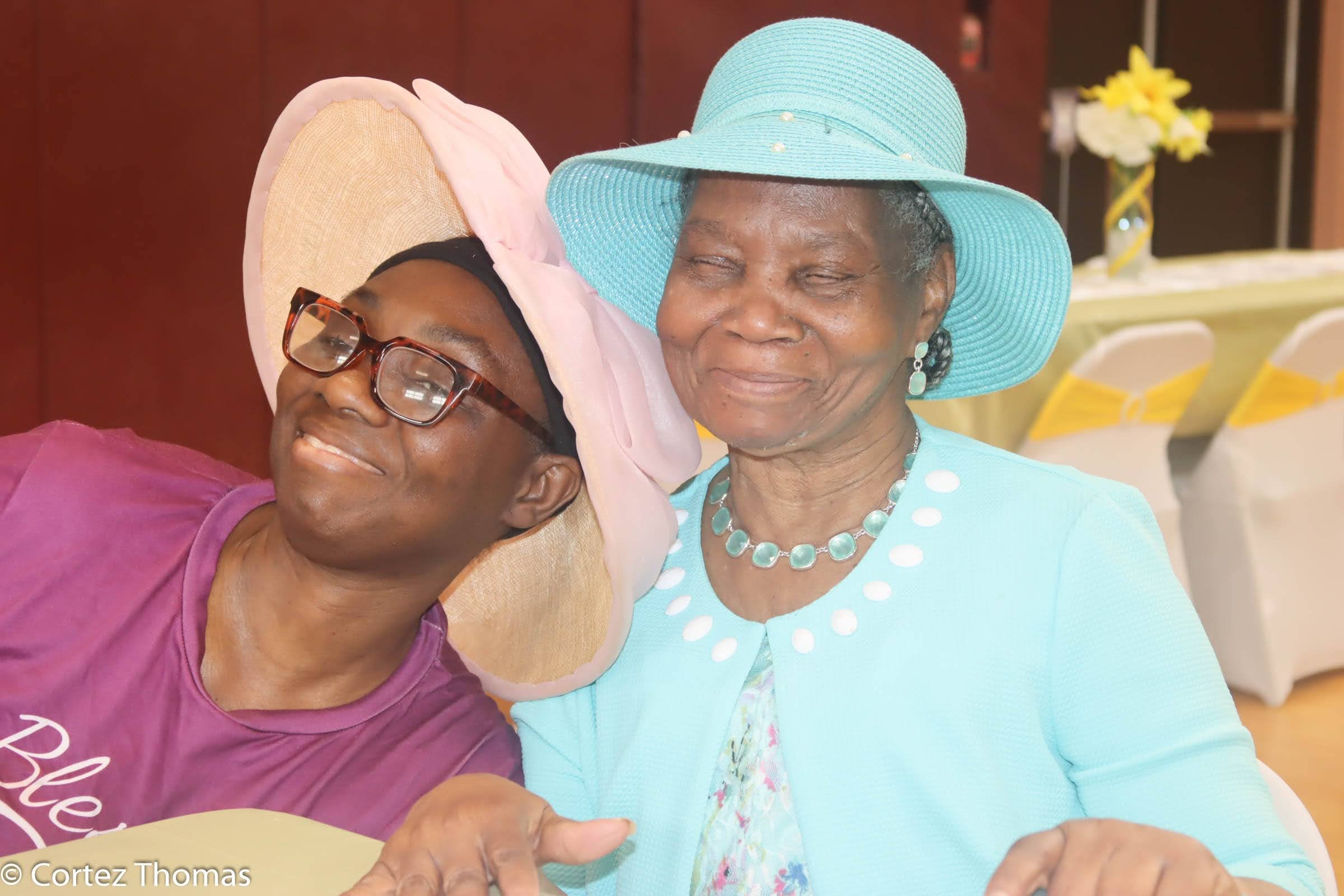 Two women, one wearing a large lavender hat and glasses and the other wearing a light blue hat and dress, sharing a joyful moment indoors.