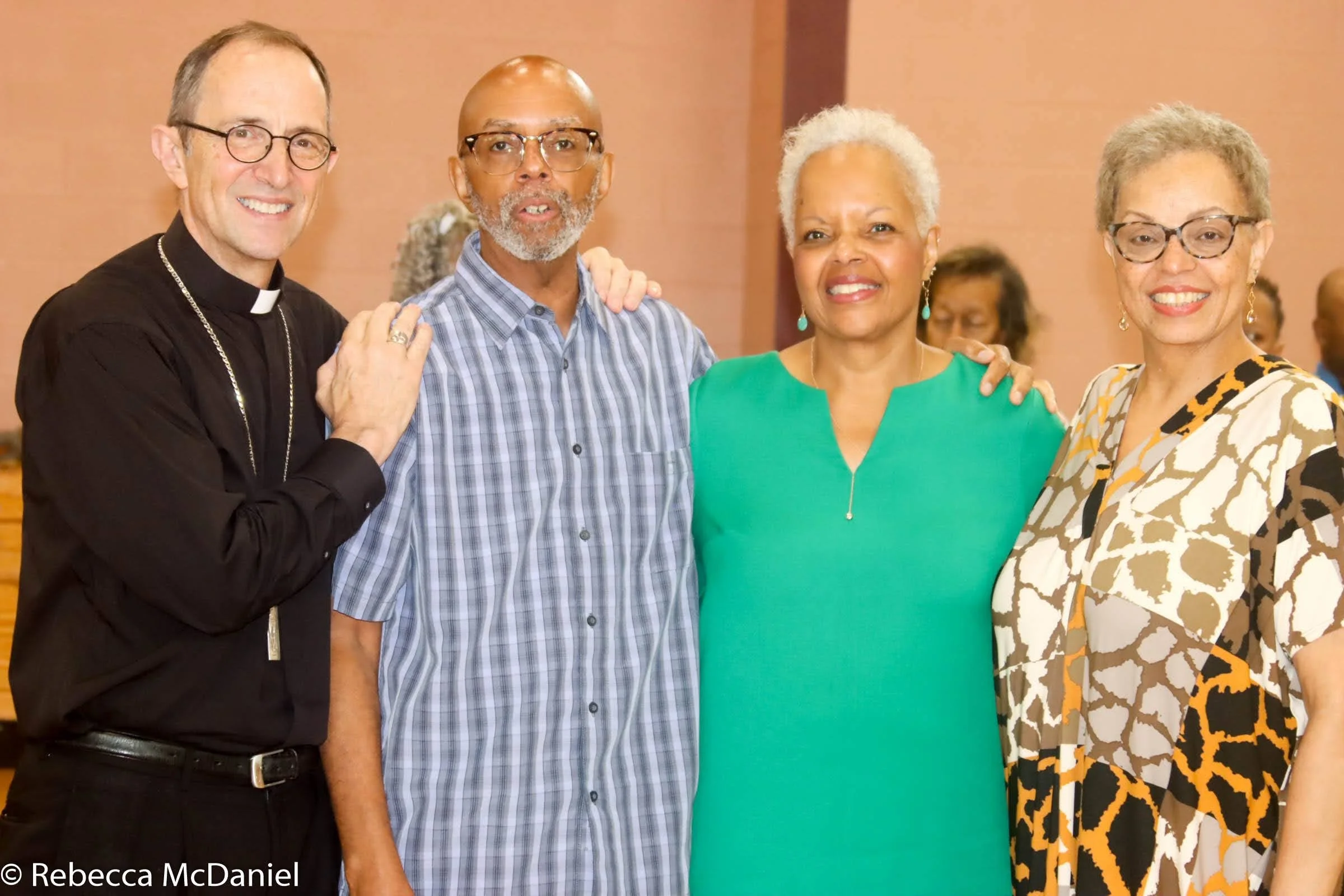 Four diverse adults standing close together at an indoor event, smiling at the camera. The man on the left is wearing clerical attire, and the other three are women wearing colorful dresses.