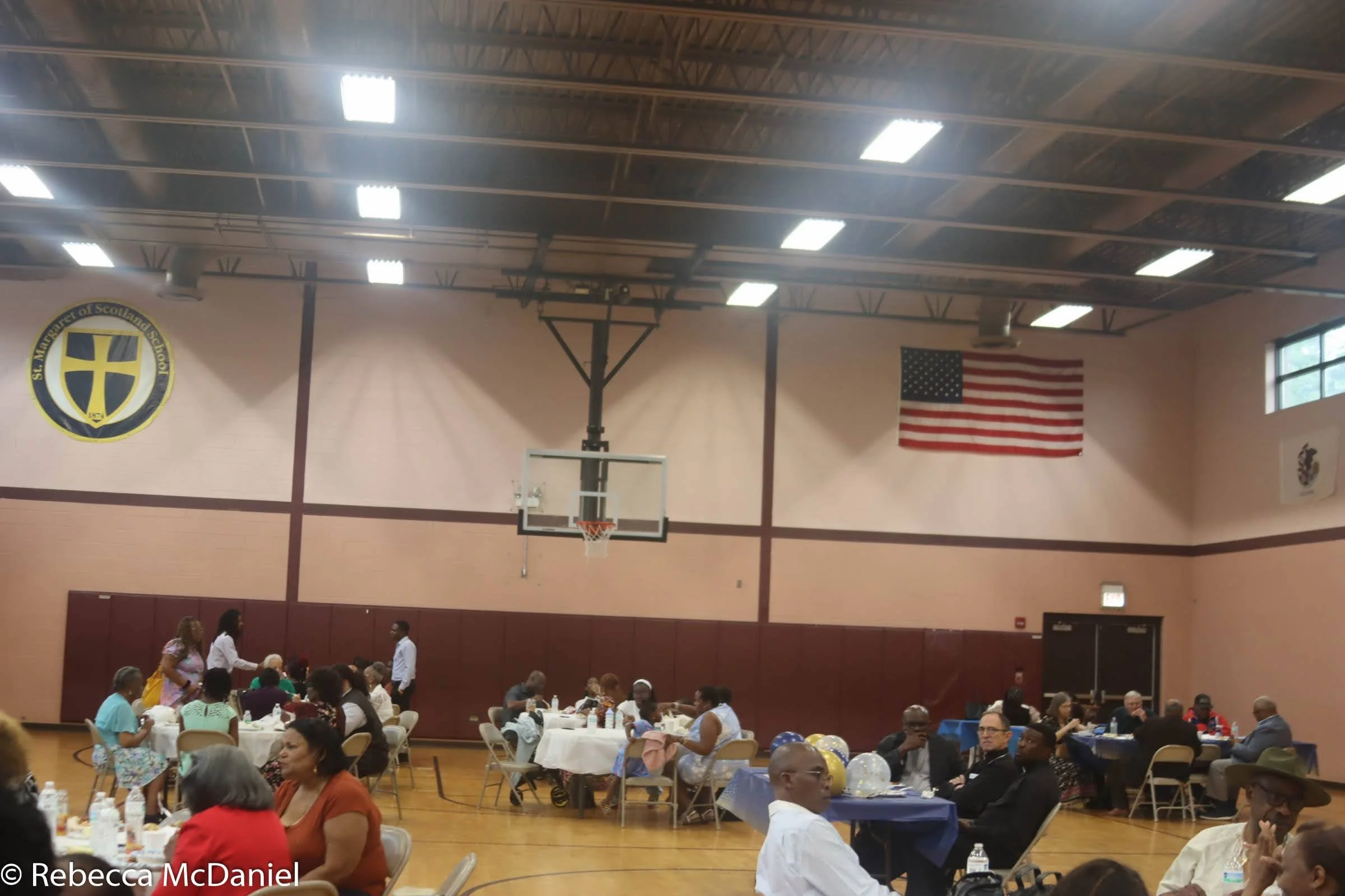 A large indoor gathering in a gymnasium with round tables and people seated, decorated with balloons, and a basketball hoop on the wall. An American flag and a school emblem are visible on the walls.