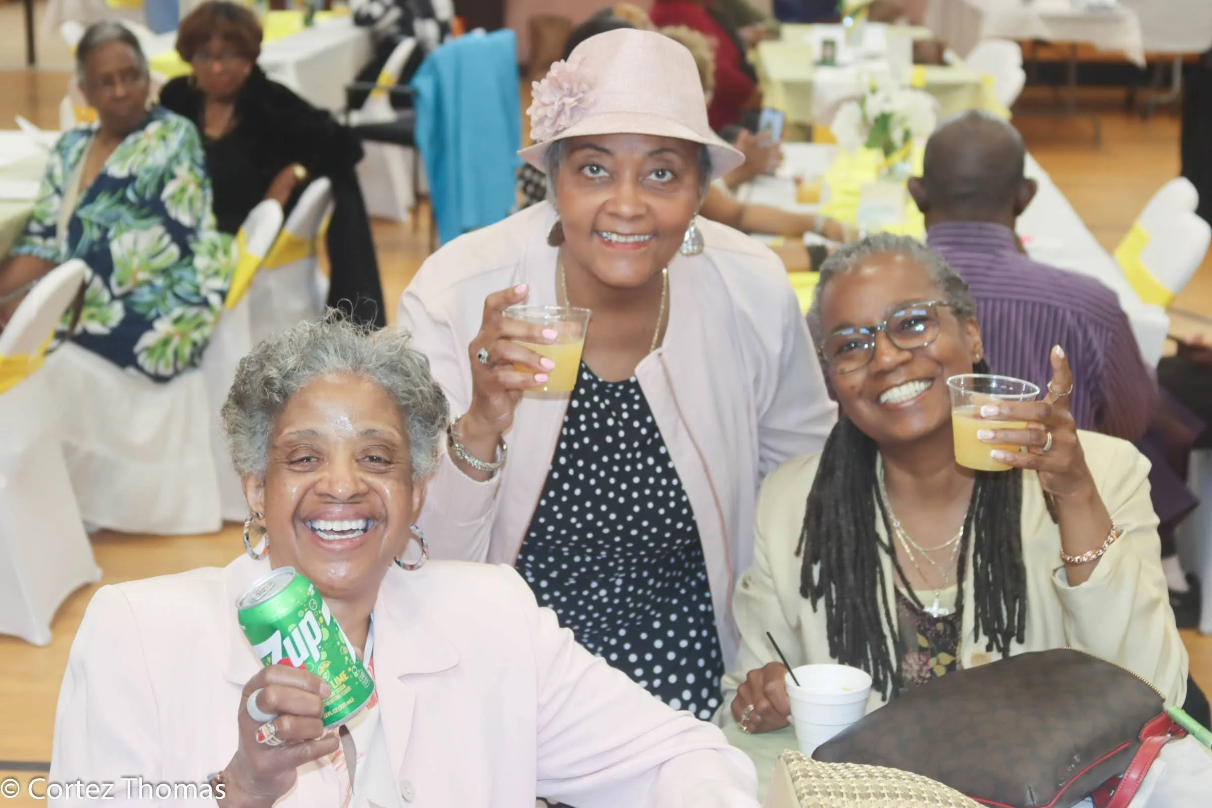 Three women at a celebration holding drinks and smiling at the camera, with a banquet hall setting in the background.