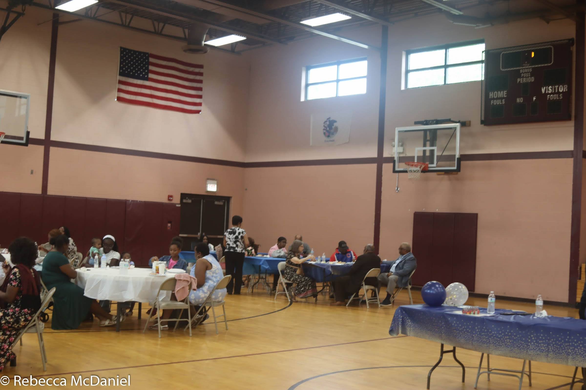 People sitting at round tables in a gymnasium decorated with blue and white balloons and tablecloths. An American flag hangs on the wall. There are basketball hoops and a scoreboard in the background.