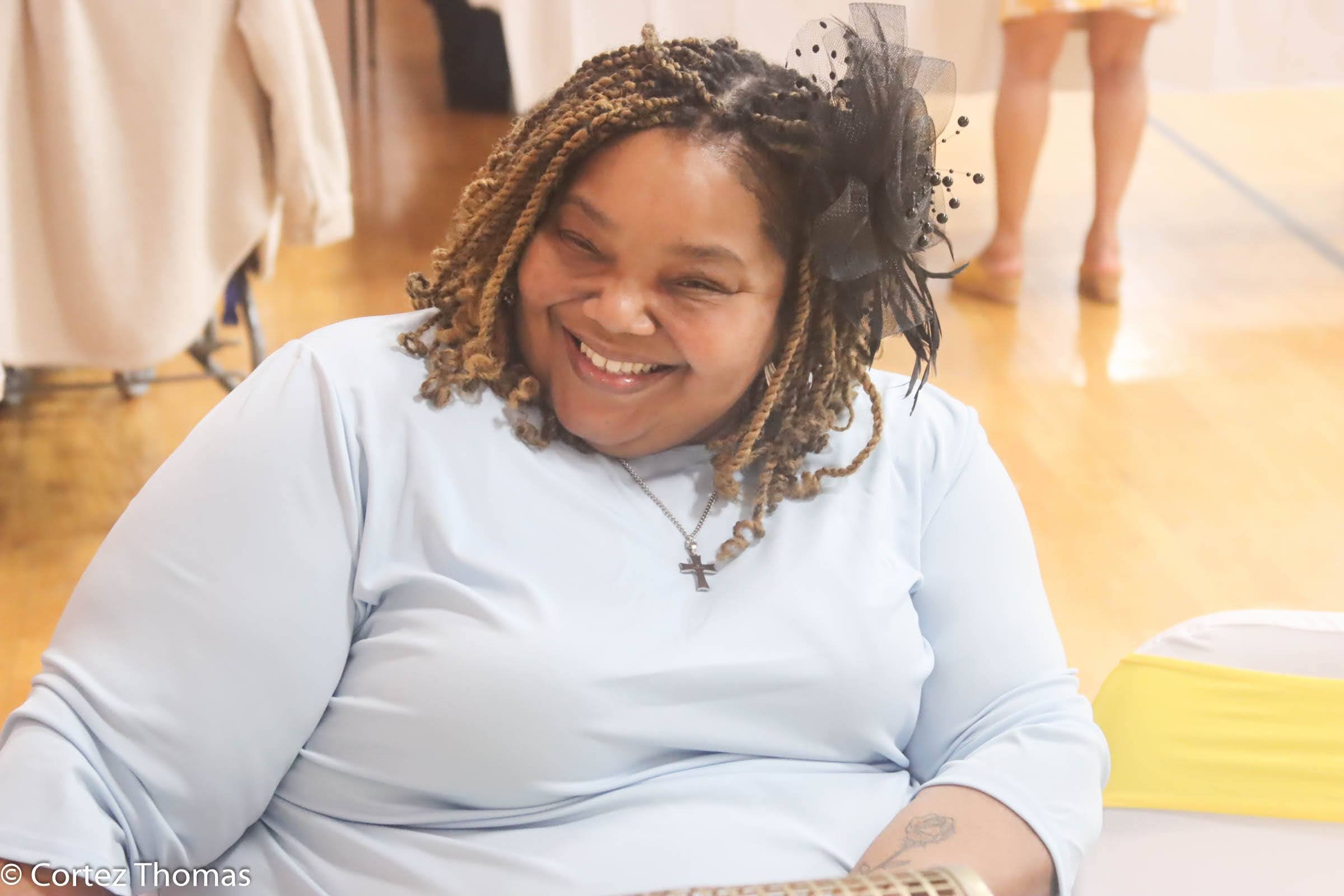 Smiling woman with tan twisted hair wearing a gray shirt, a cross necklace, and a black festive headband, seated indoors.