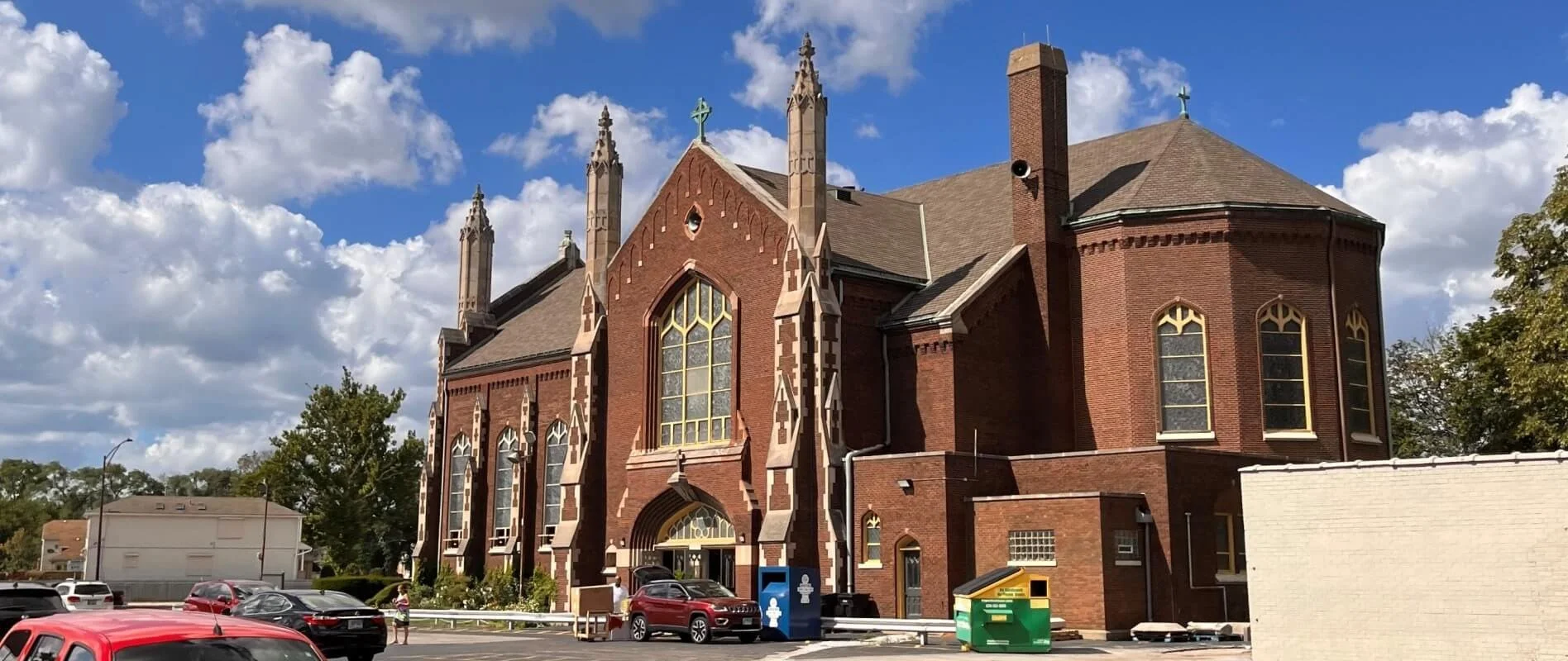A large red brick church with Gothic architectural features, pointed arch windows with yellow trim, and ornate spires on the roof. It is situated in a parking lot with several cars and a few people nearby. The sky above is partly cloudy with blue and