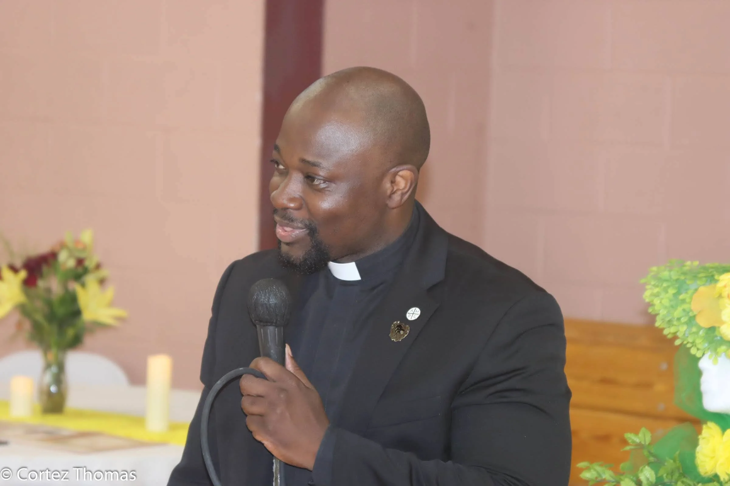 Man in black clergy suit holding microphone, speaking at an indoor event with flowers and candles in the background.
