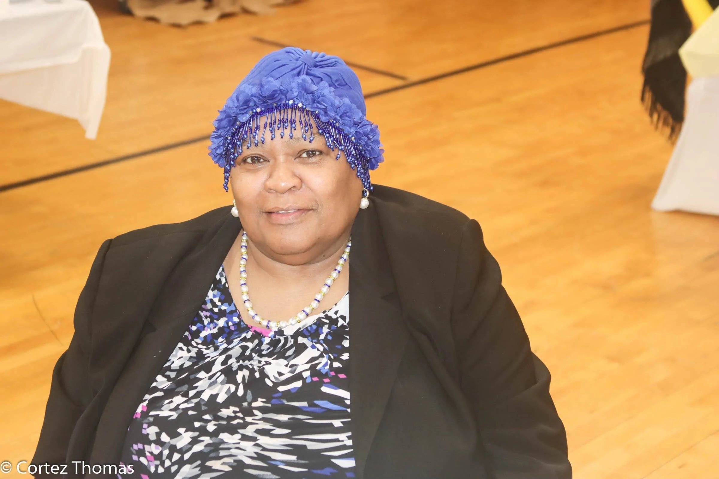 A woman wearing a bright blue headwrap and beaded jewelry, sitting indoors on a wooden floor with decorated tables in the background.