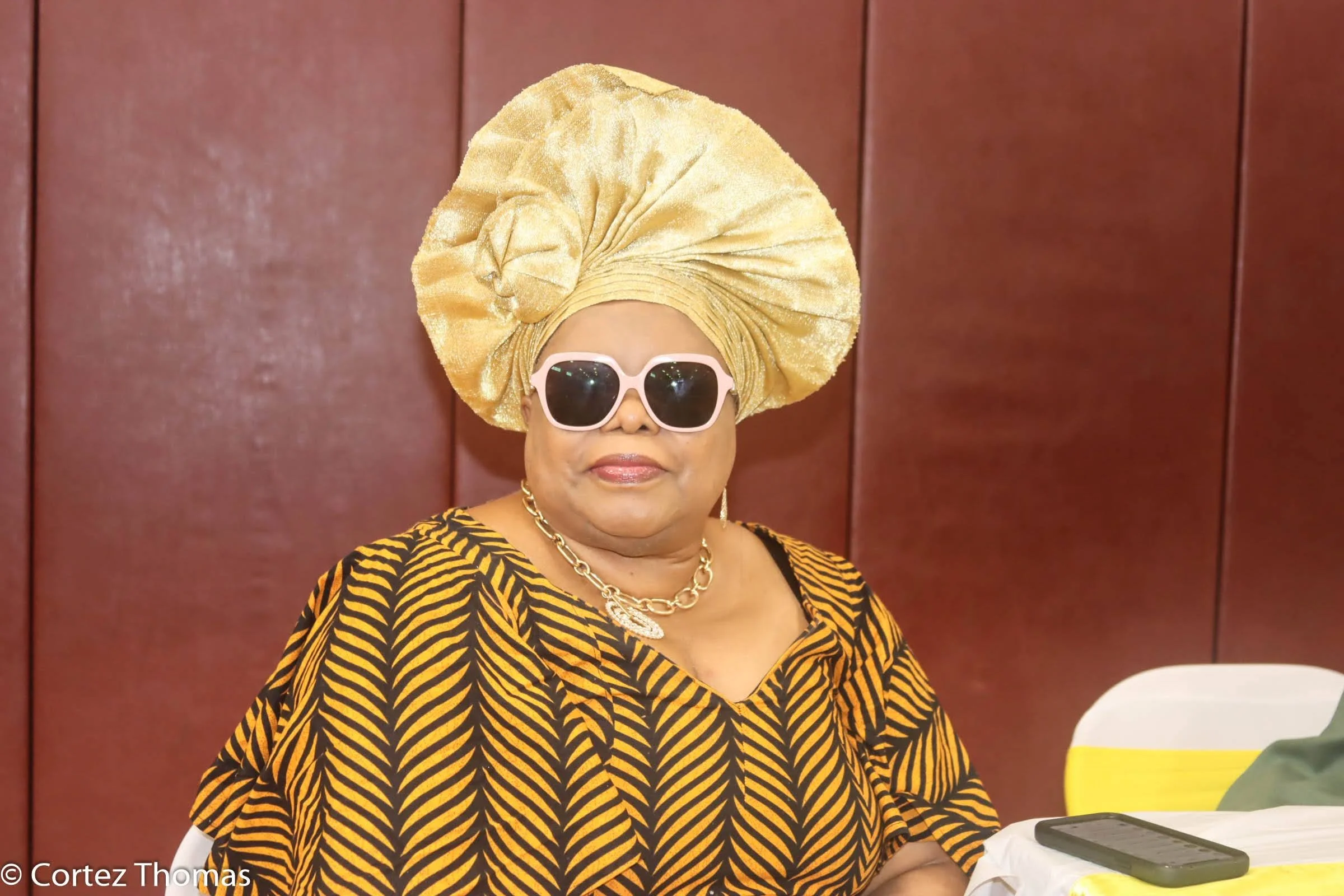 Woman wearing a large gold headwrap, pink sunglasses, gold jewelry, and a black and yellow patterned dress, sitting at a table with a phone and papers, against a brown backdrop.