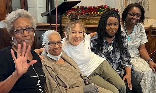 Five women sitting together indoors, smiling, with a piano in the background.