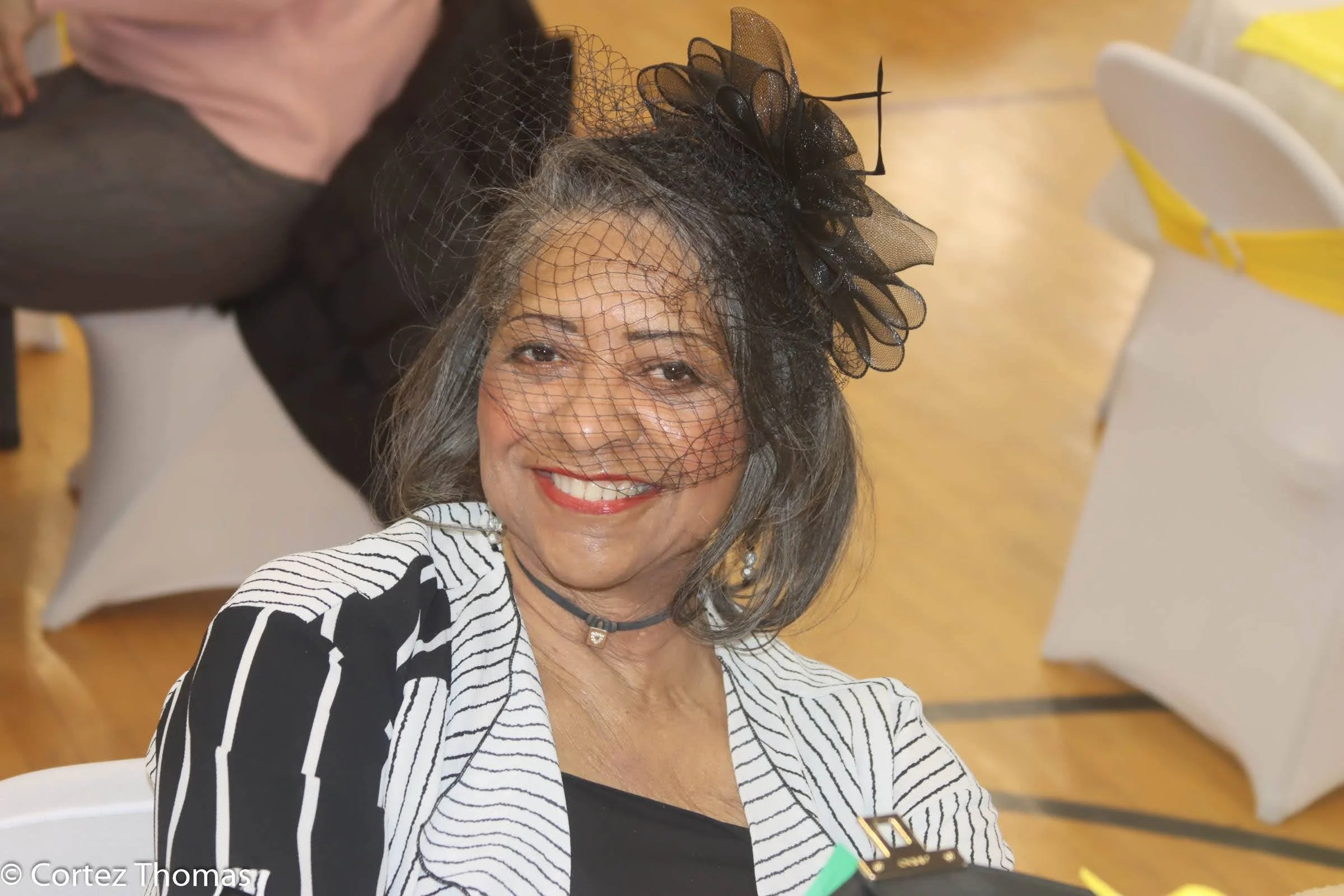 Smiling woman with gray hair wearing a black and white striped blazer and black top, accessorized with a black choker and rhinestone earrings, is wearing a black fascinator with mesh netting covering her face, at a social event or celebration.