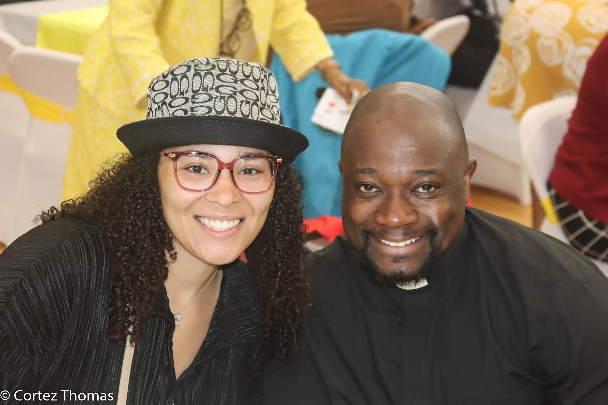 A smiling woman with glasses and a hat posing next to a smiling priest in a black shirt.