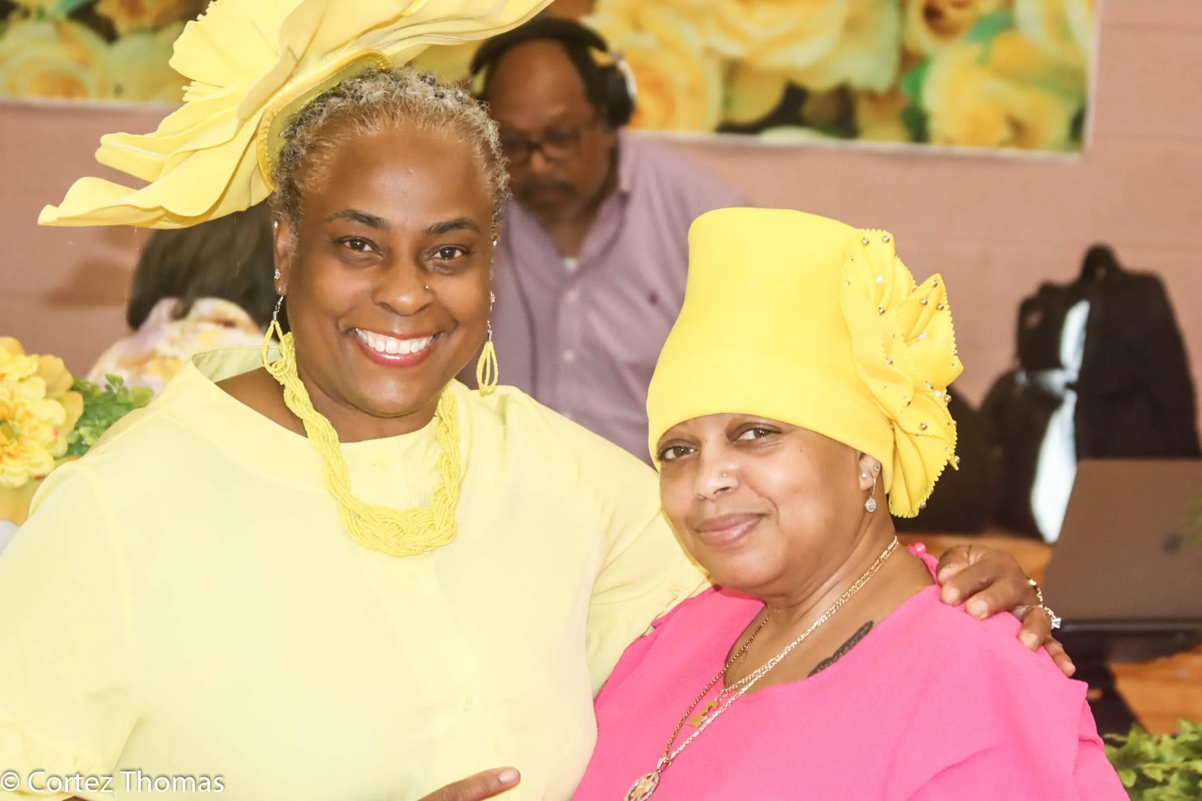 Two women smiling and posing together, dressed in bright yellow and pink traditional clothing, wearing elaborate headwraps, with a man in the background wearing headphones and glasses. The setting appears to be a celebration or gathering with floral 