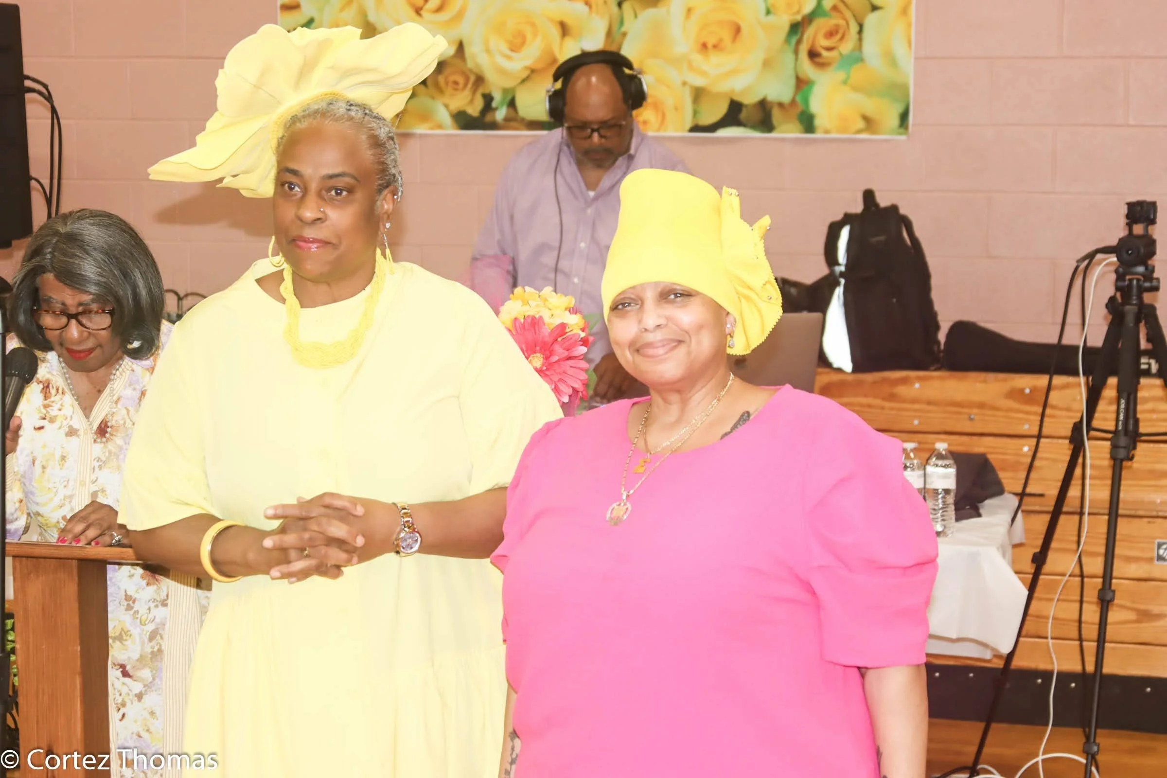 Two women in bright dresses shaking hands at a celebration or event, with a woman speaking into a microphone and a man with headphones in the background.