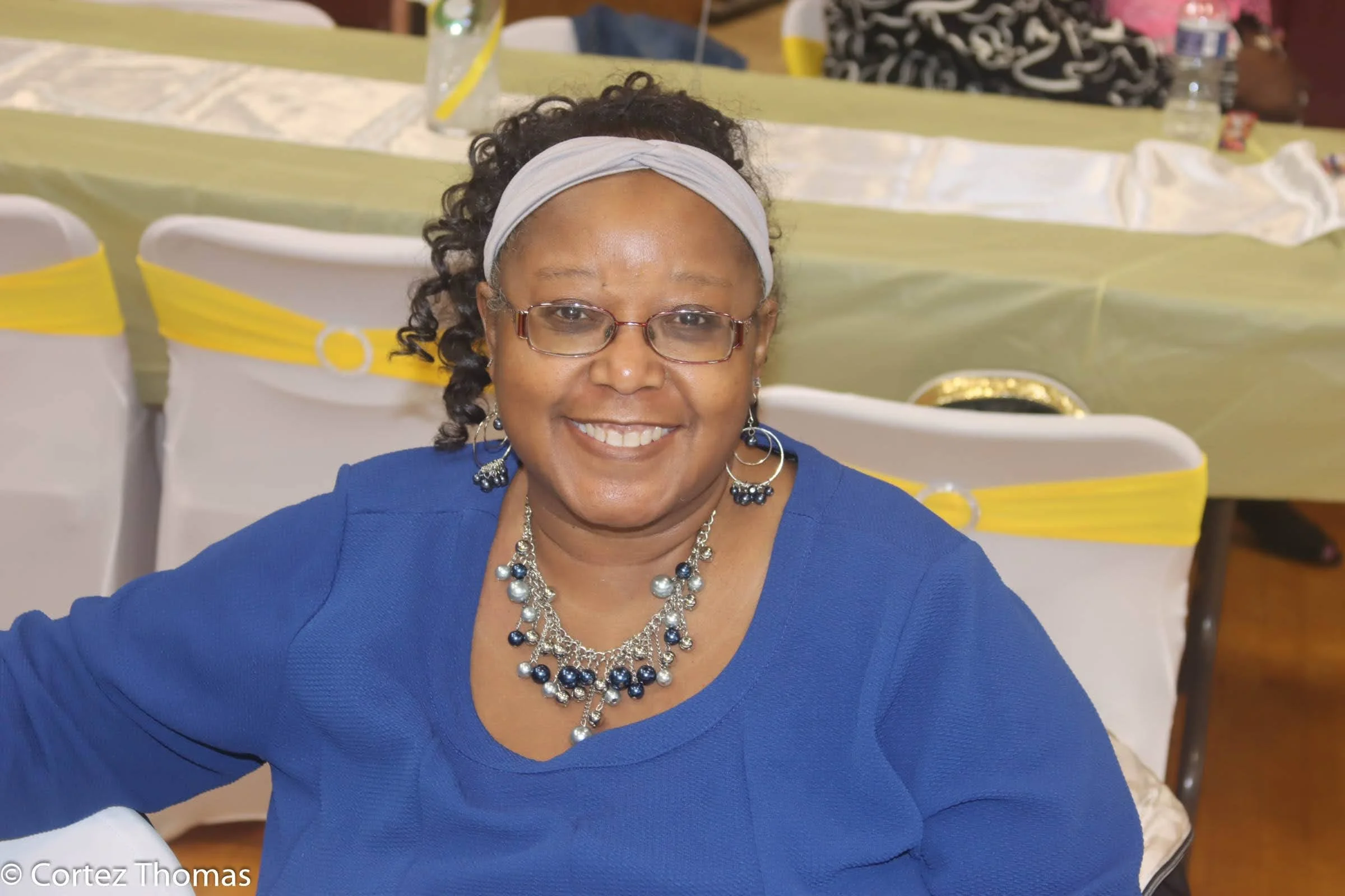 A smiling woman with glasses and earrings, wearing a blue top and a white headband, sitting at a decorated event table.