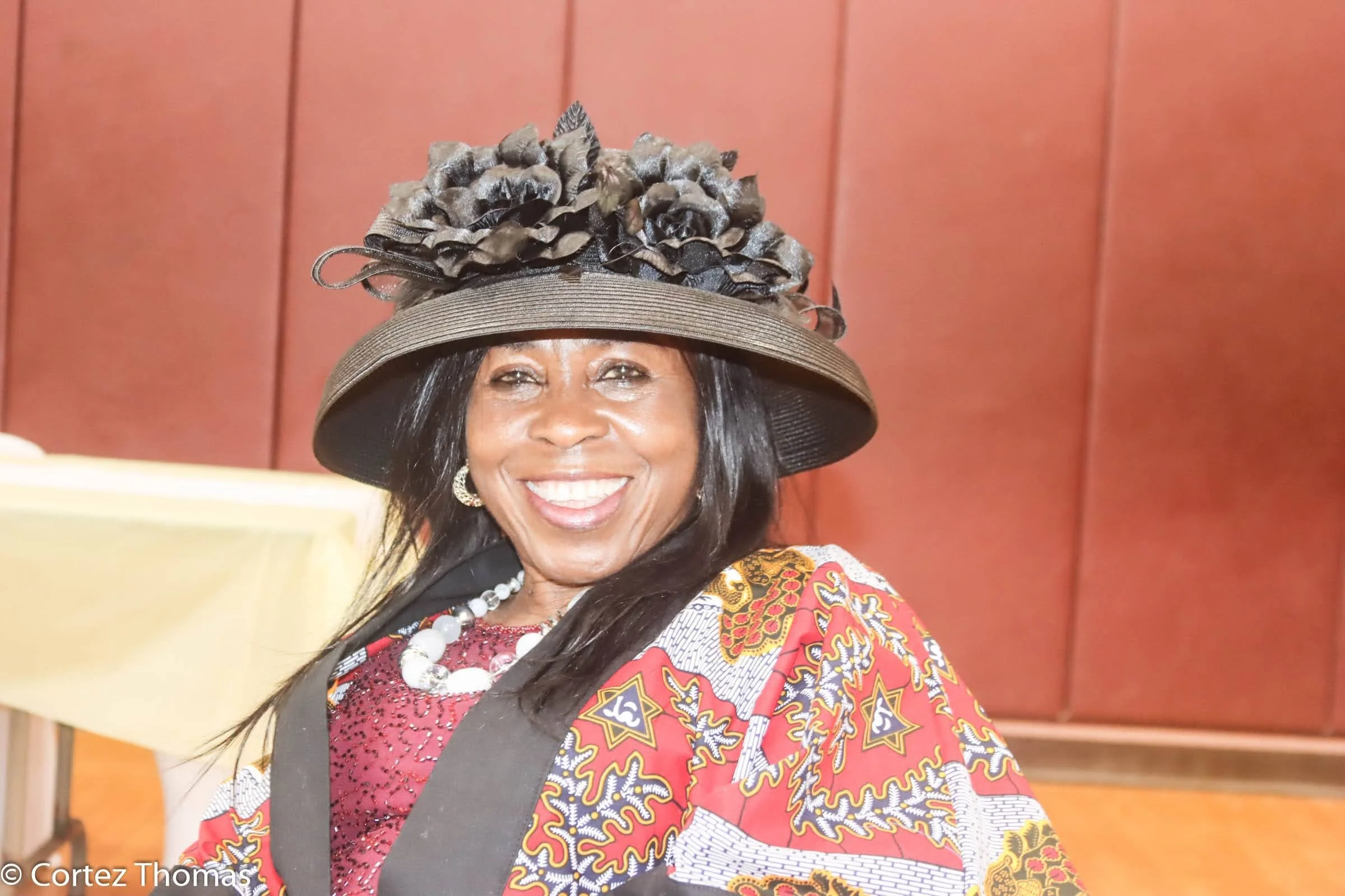 A smiling woman wearing a large, decorative hat with floral accents, black and in a neutral tone, a colorful patterned outfit, pearl earrings, and a pearl necklace, in front of a wood-paneled background.