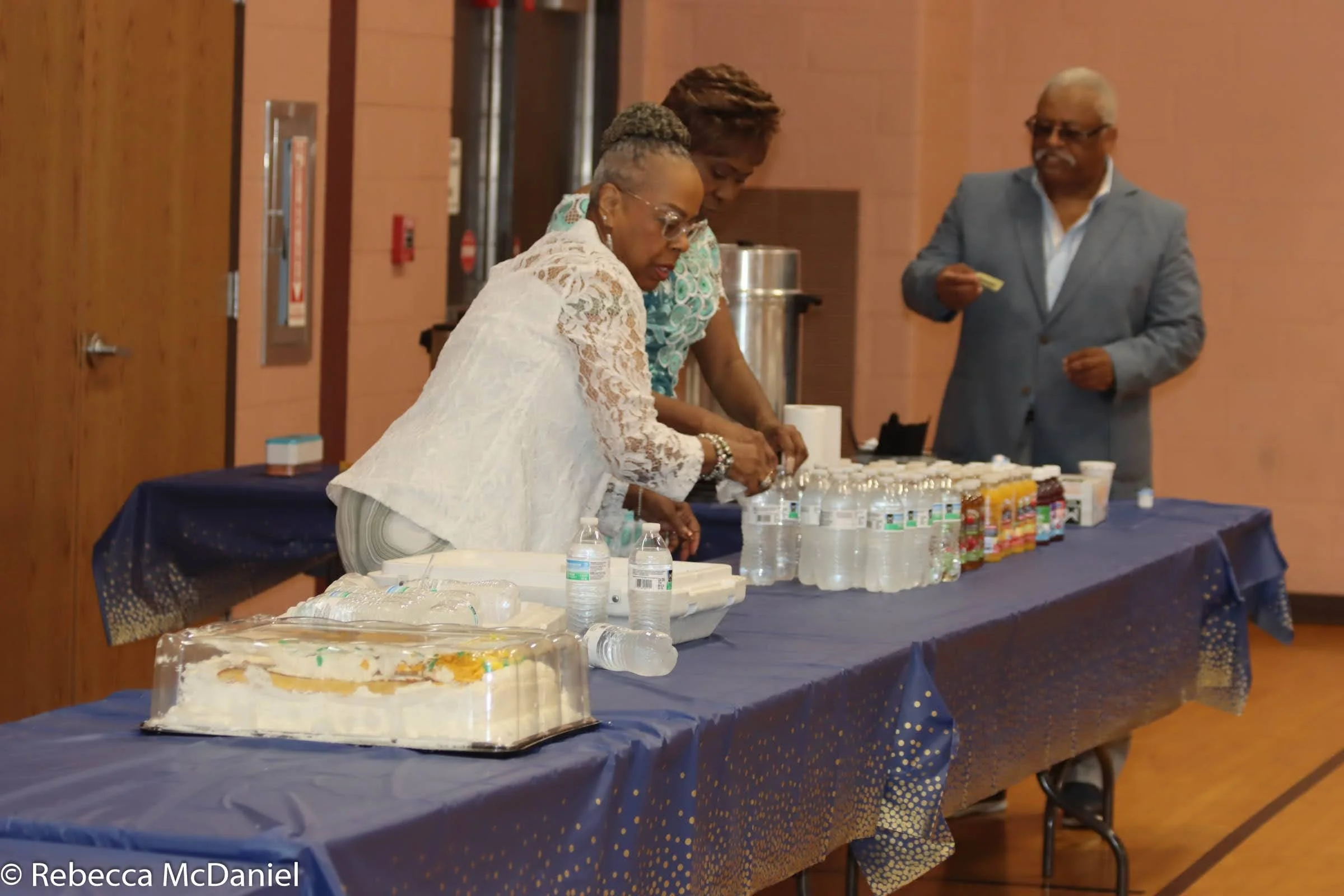 Three people stand at a table with bottled water, snacks, and a cake in a room with warm-colored walls and wooden doors.