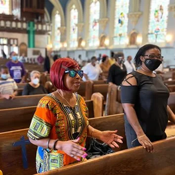 Two women with face masks inside a church, one with red hair and colorful dress and the other with black hair and black top, surrounded by people seated in pews.