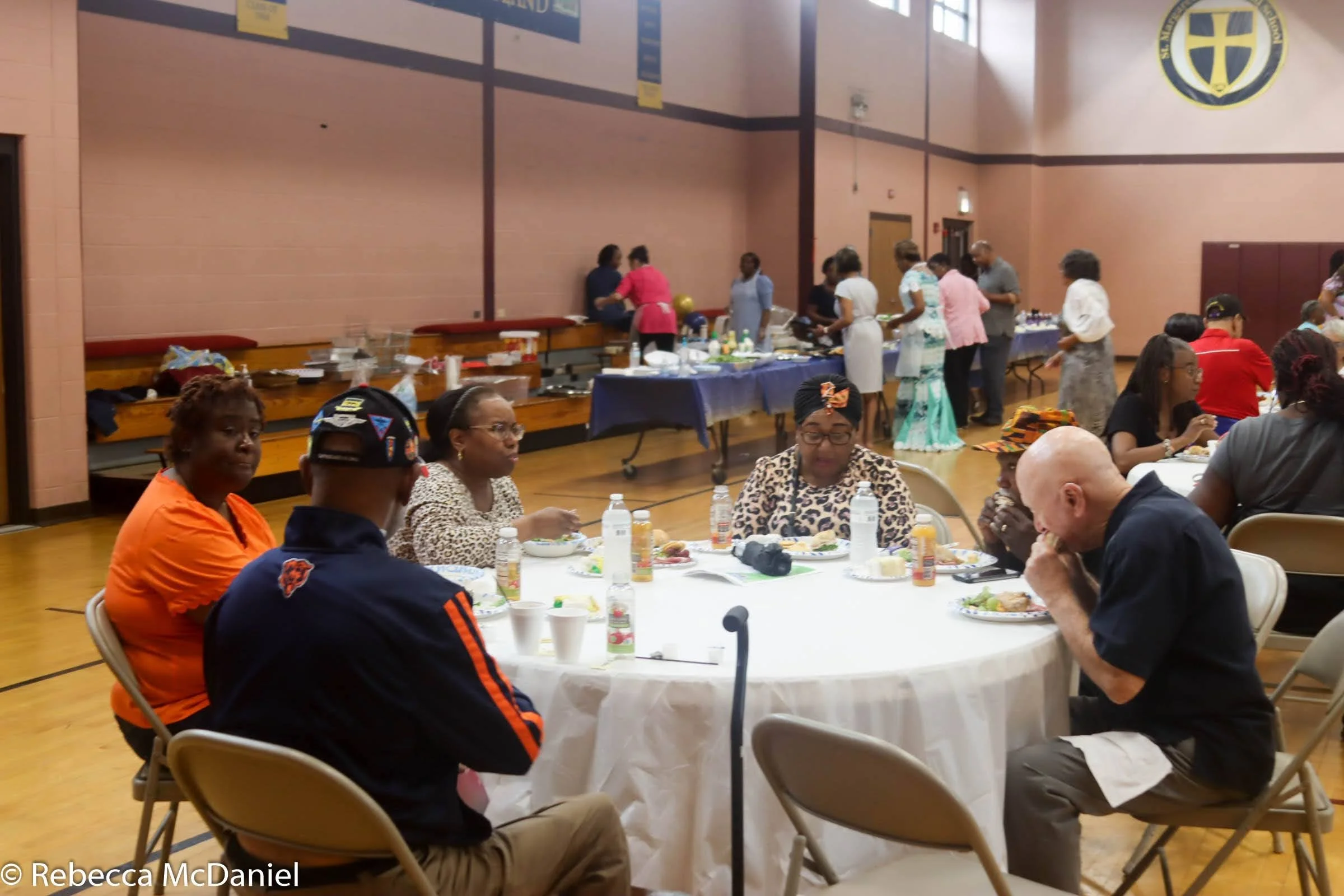 People sitting around a round table eating at an indoor event with food and drinks, with a buffet table in the background and others preparing food.