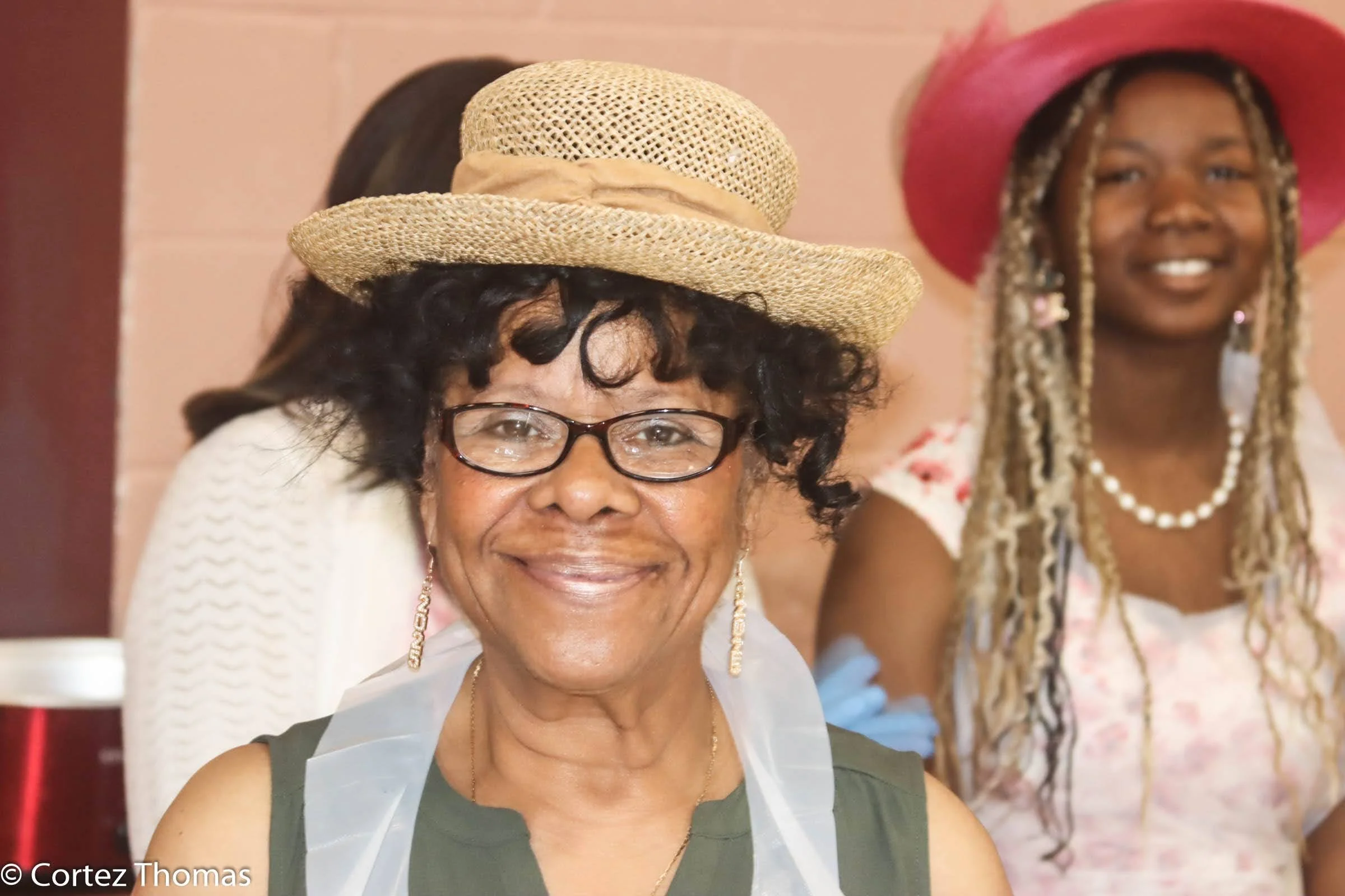Smiling woman wearing glasses, a straw hat, and earrings, with a young girl wearing a pink hat, pearl necklace, and floral dress in the background.