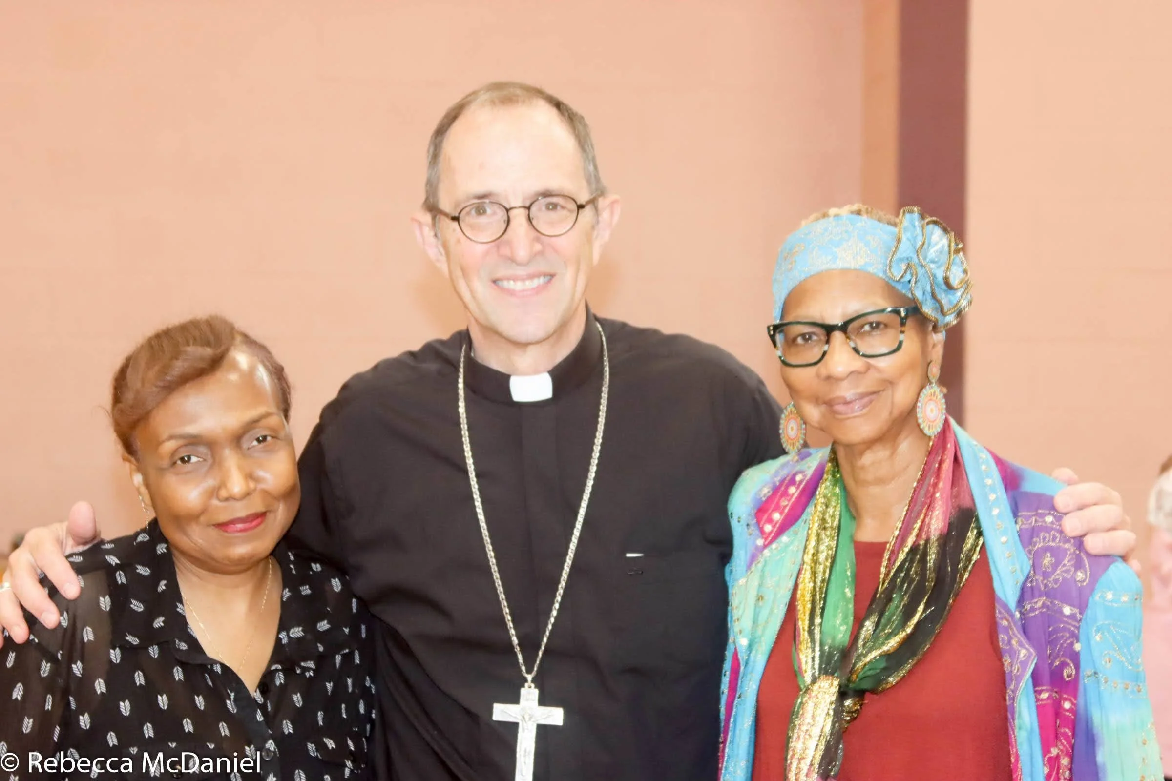 Three smiling women and a man, with the man dressed as a priest, standing together indoors.