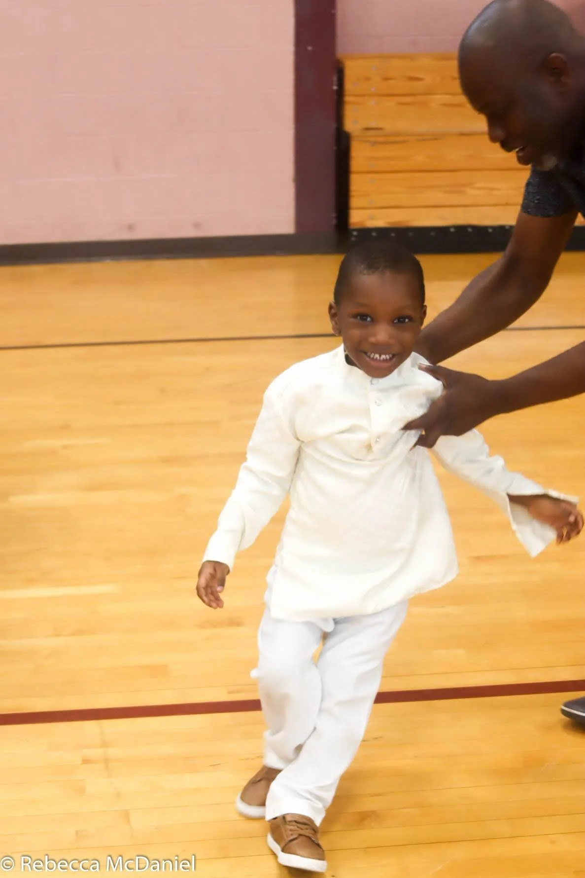 A young boy dressed in white clothing, smiling and spinning around on a wooden floor, with an adult man holding his waist and supporting him.