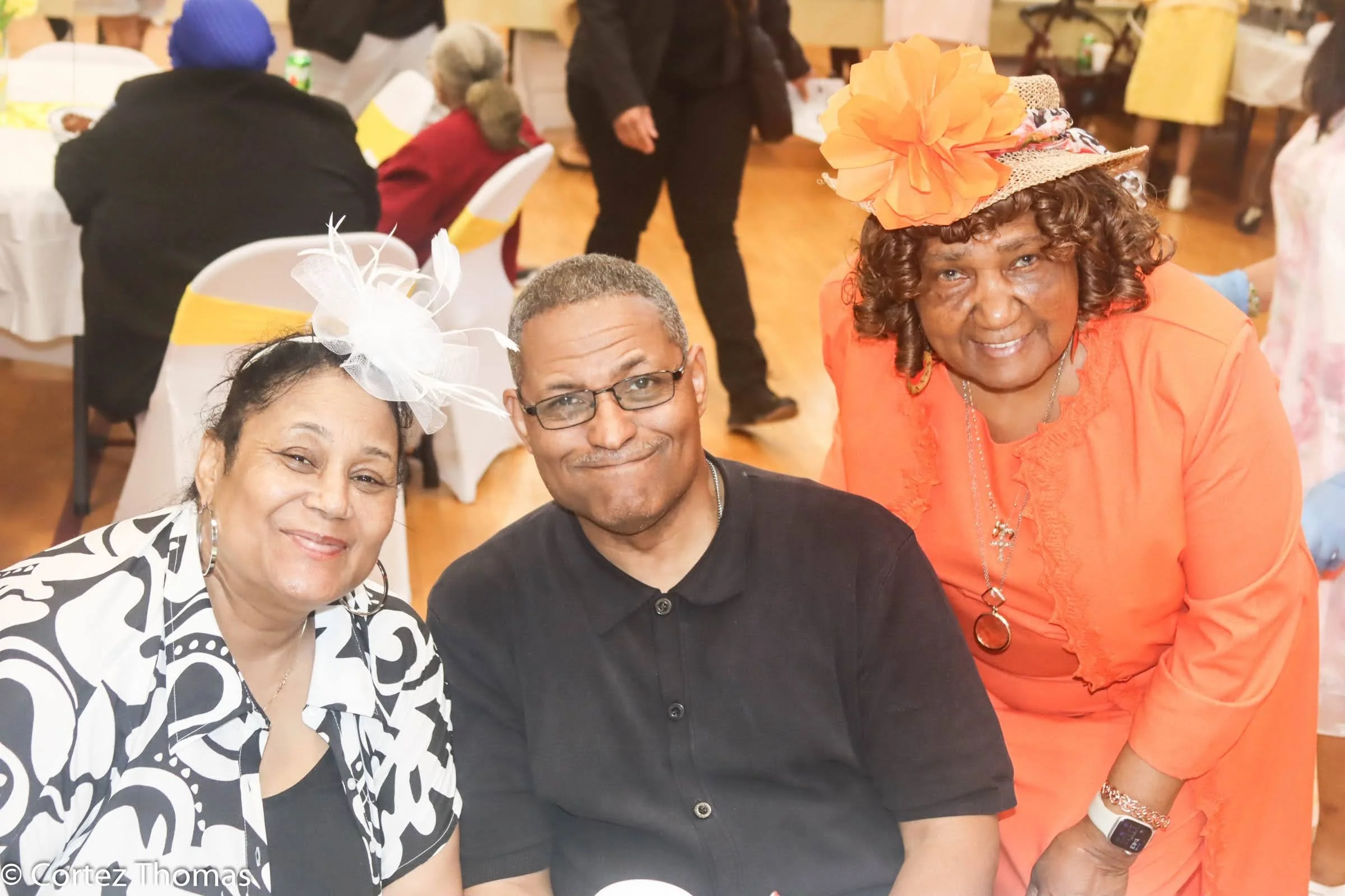 Three adults sitting together at a party or celebration, smiling and wearing festive attire, with round dining tables and chairs in the background.