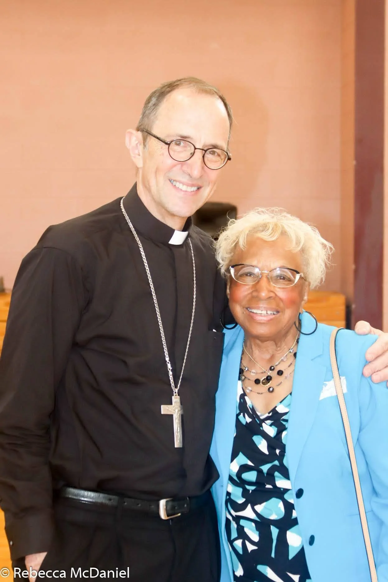 A smiling church clergyman wearing a black clergy shirt with a white collar, glasses, and a silver cross necklace, standing next to an older woman with short curly blonde hair, glasses, and colorful attire, both posing happily together.