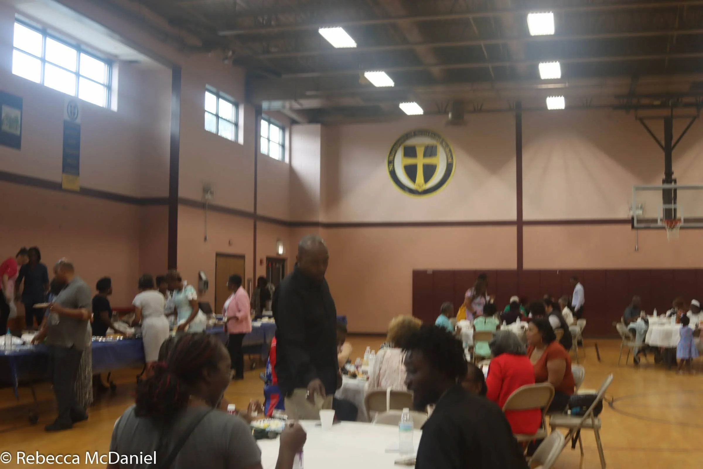 People gathered in a gymnasium for a social event, with tables set up for food and drinks, a basketball hoop, and a large emblem on the wall.