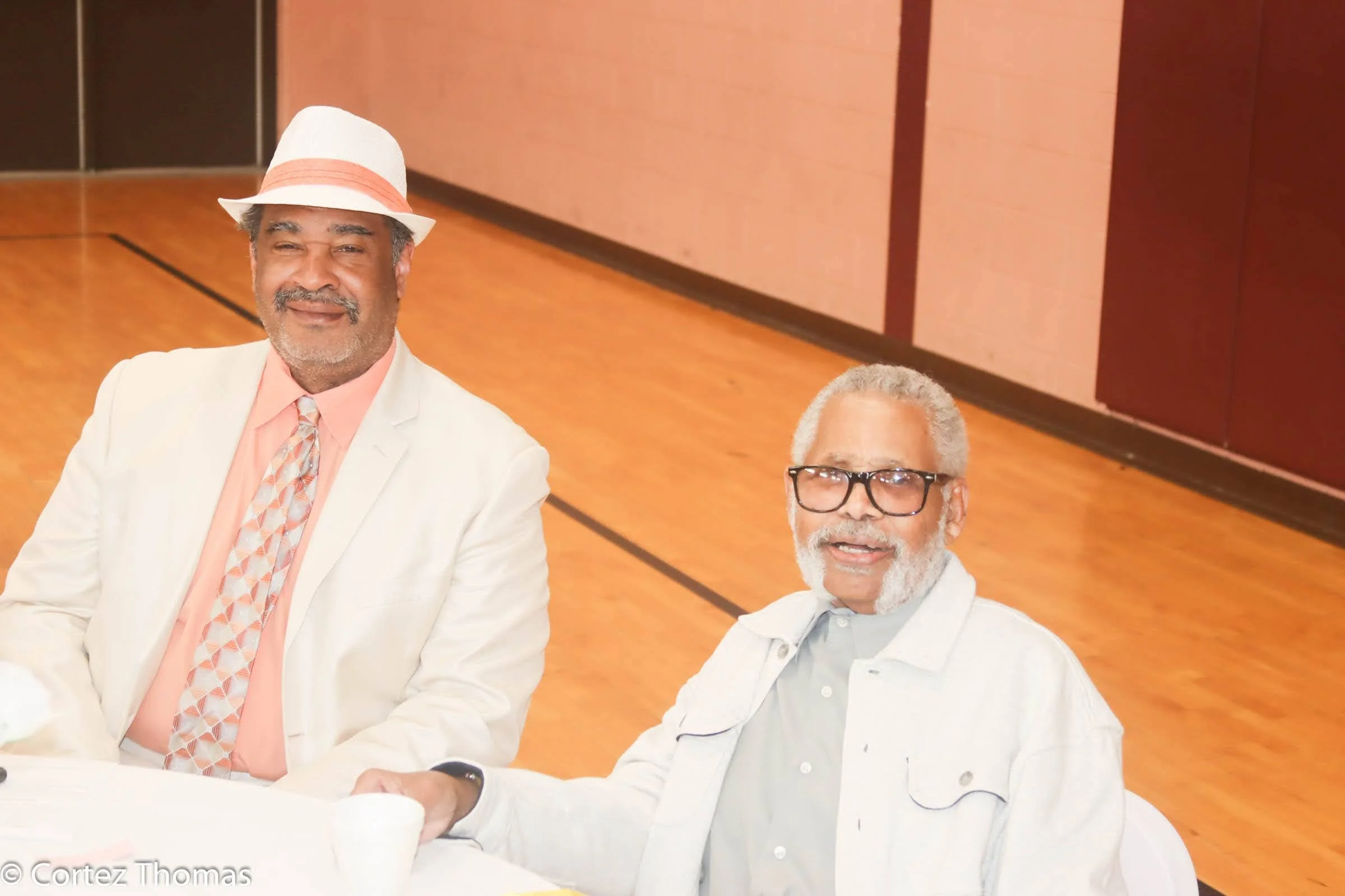 Two elderly men sitting at a table, smiling, one wearing a white suit, peach shirt, patterned tie, white hat with a peach band, and the other wearing glasses, white jacket, and light grey shirt, in a wooden-floored room.
