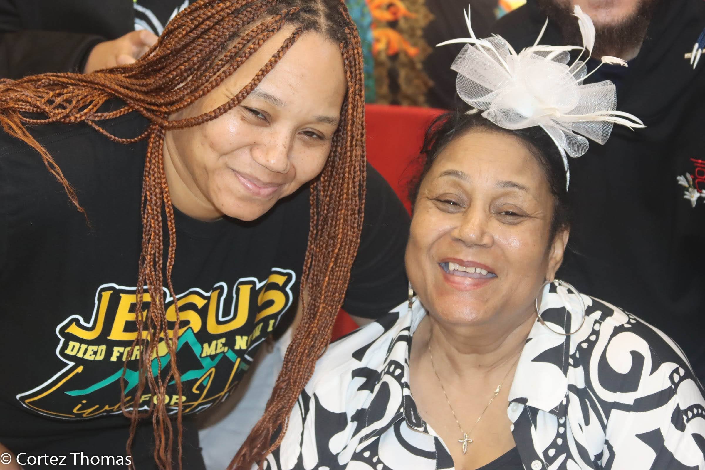 Two women smiling, one with braided hair and wearing a black T-shirt, the other with a colorful hair accessory and wearing a black and white patterned shirt.