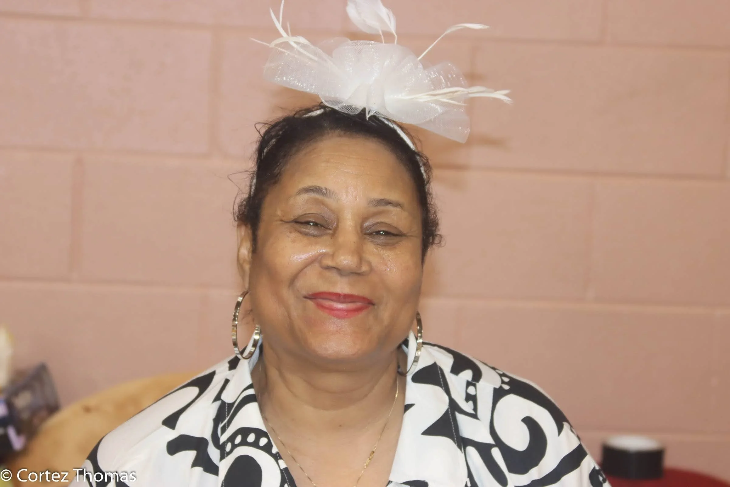 A smiling middle-aged woman wearing a large white decorative headpiece and a black-and-white patterned shirt, standing in front of a pink brick wall.
