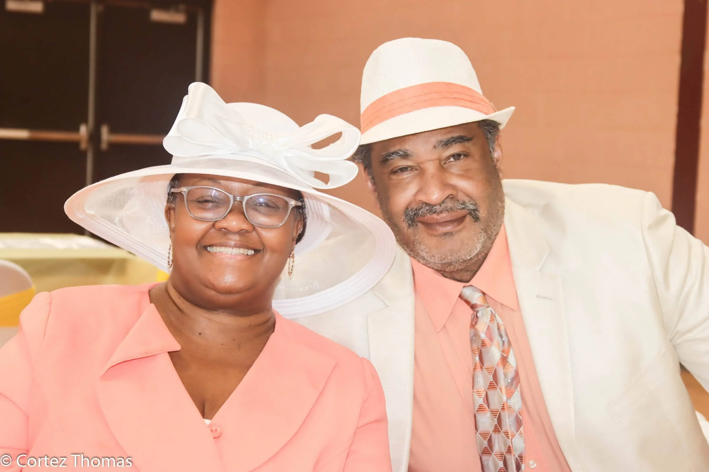 Smiling woman and man dressed in light-colored suits and stylish hats at a social event.