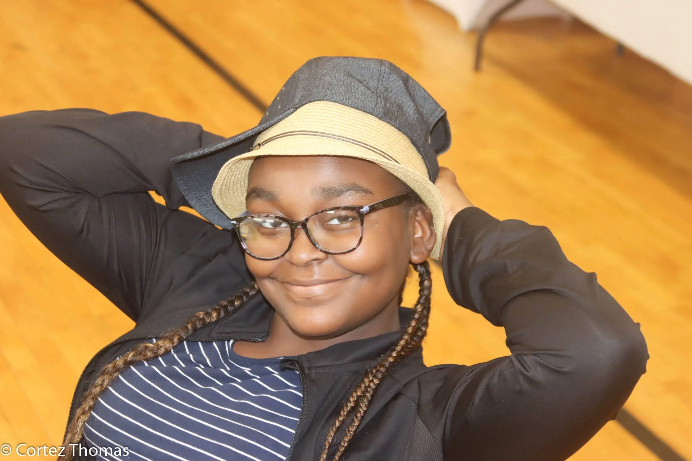 A young person smiling, wearing glasses, a striped shirt, a black jacket, and a beige fedora hat, with a wooden floor background.