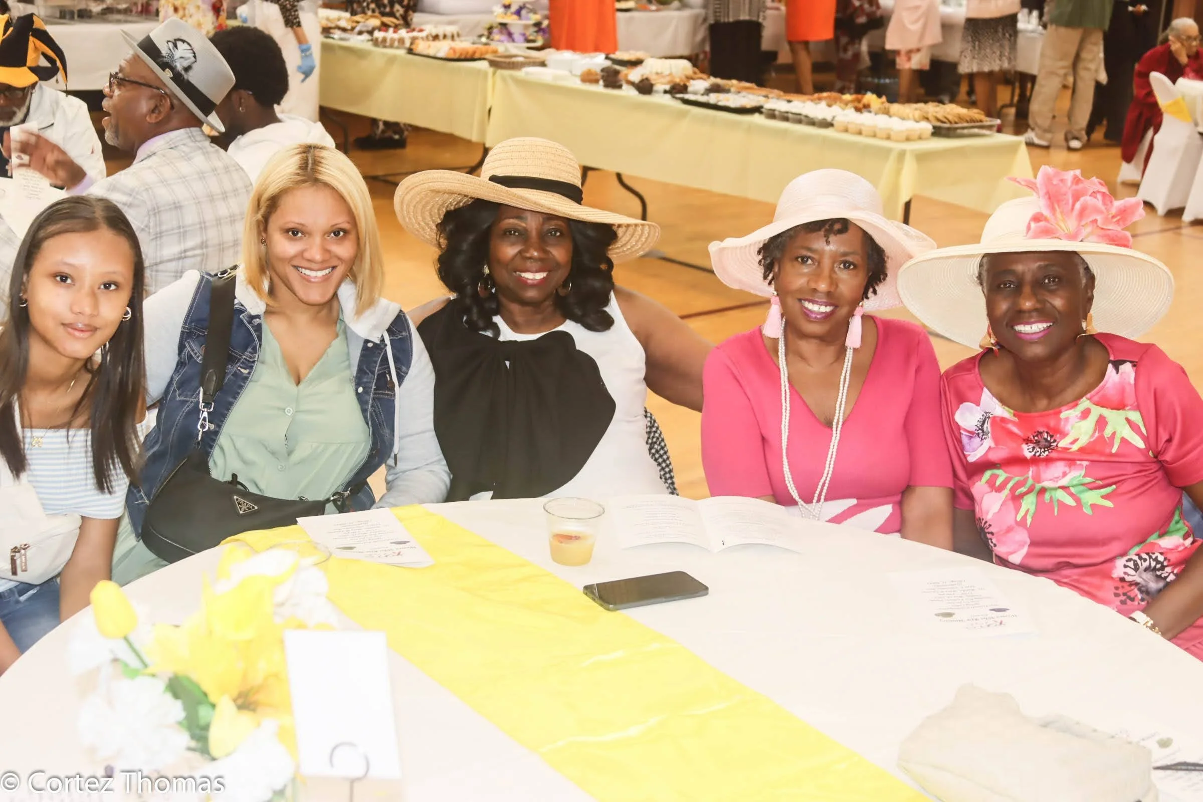 Five women sitting at a round table, smiling at the camera, during a social event or celebration. They are wearing colorful clothing and large hats. In the background, there are tables with food and other guests.
