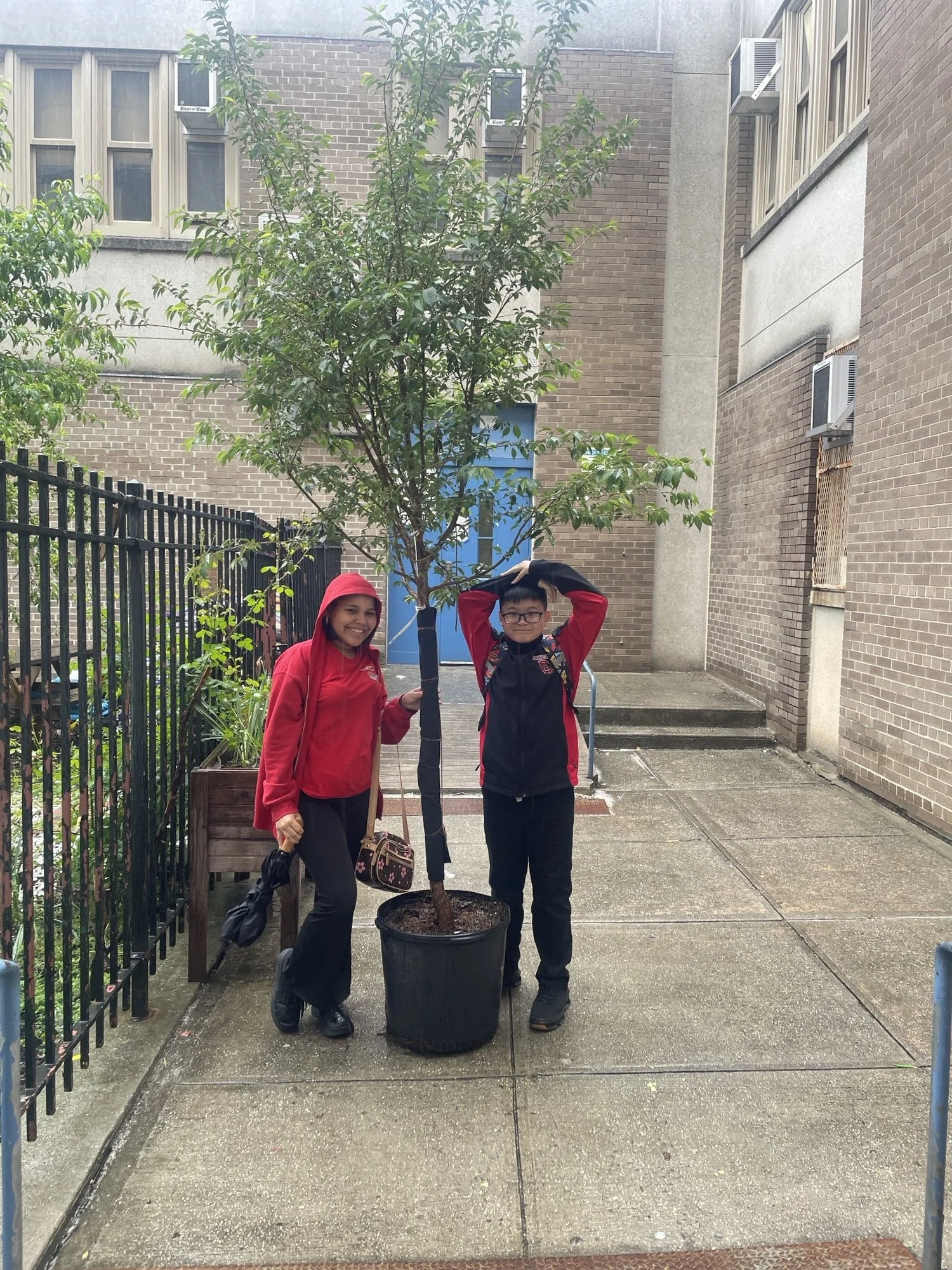 Two children, a girl and a boy, standing next to a potted tree outdoors, smiling at the camera. The girl is wearing a red jacket with a hood, and the boy is wearing glasses and a black jacket with red accents. The background includes brick buildings, windows, and steps.