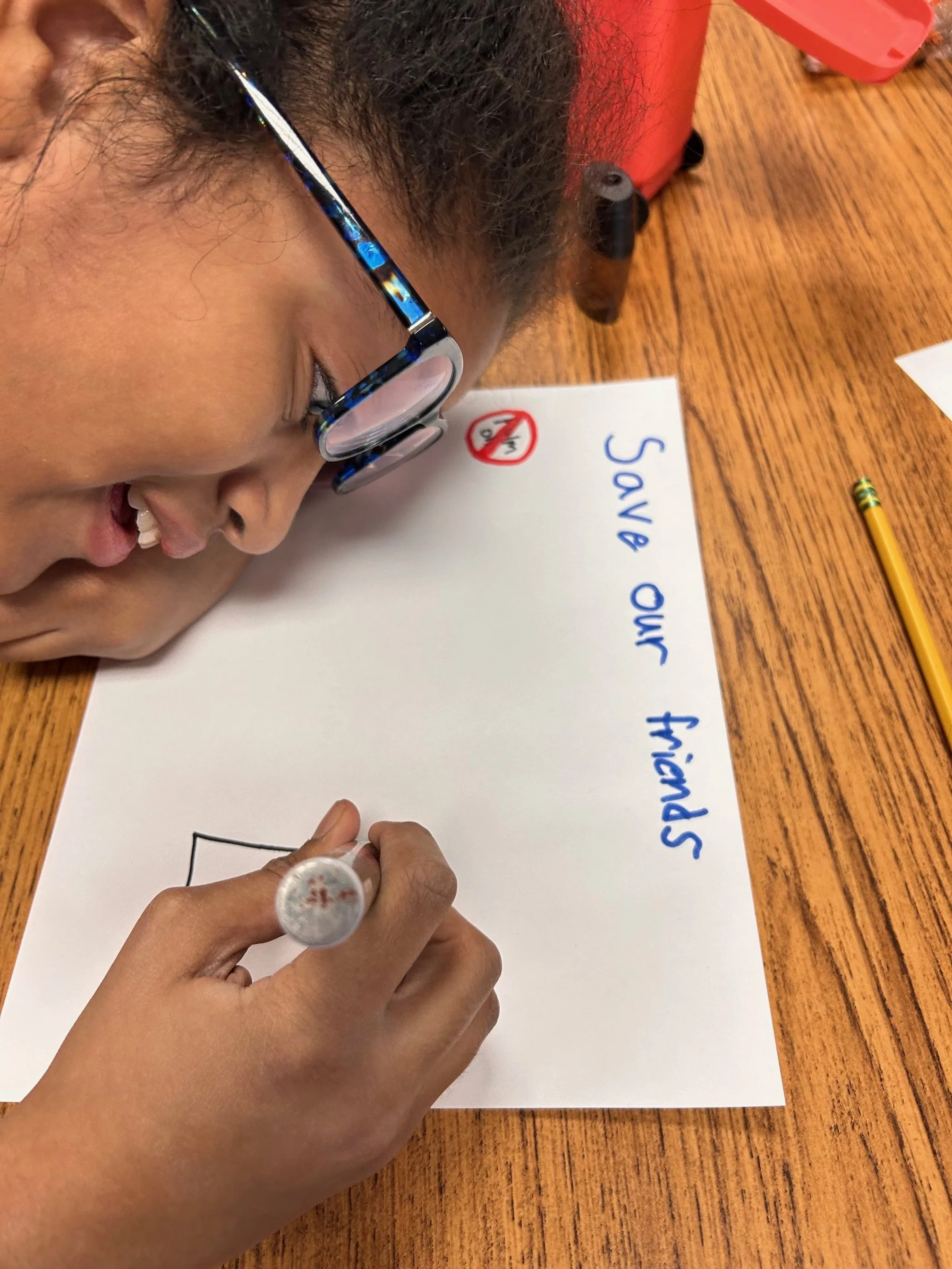 Child writing a message on a white sheet of paper with a black marker, saying 'Save Our Schools', on a wooden table.