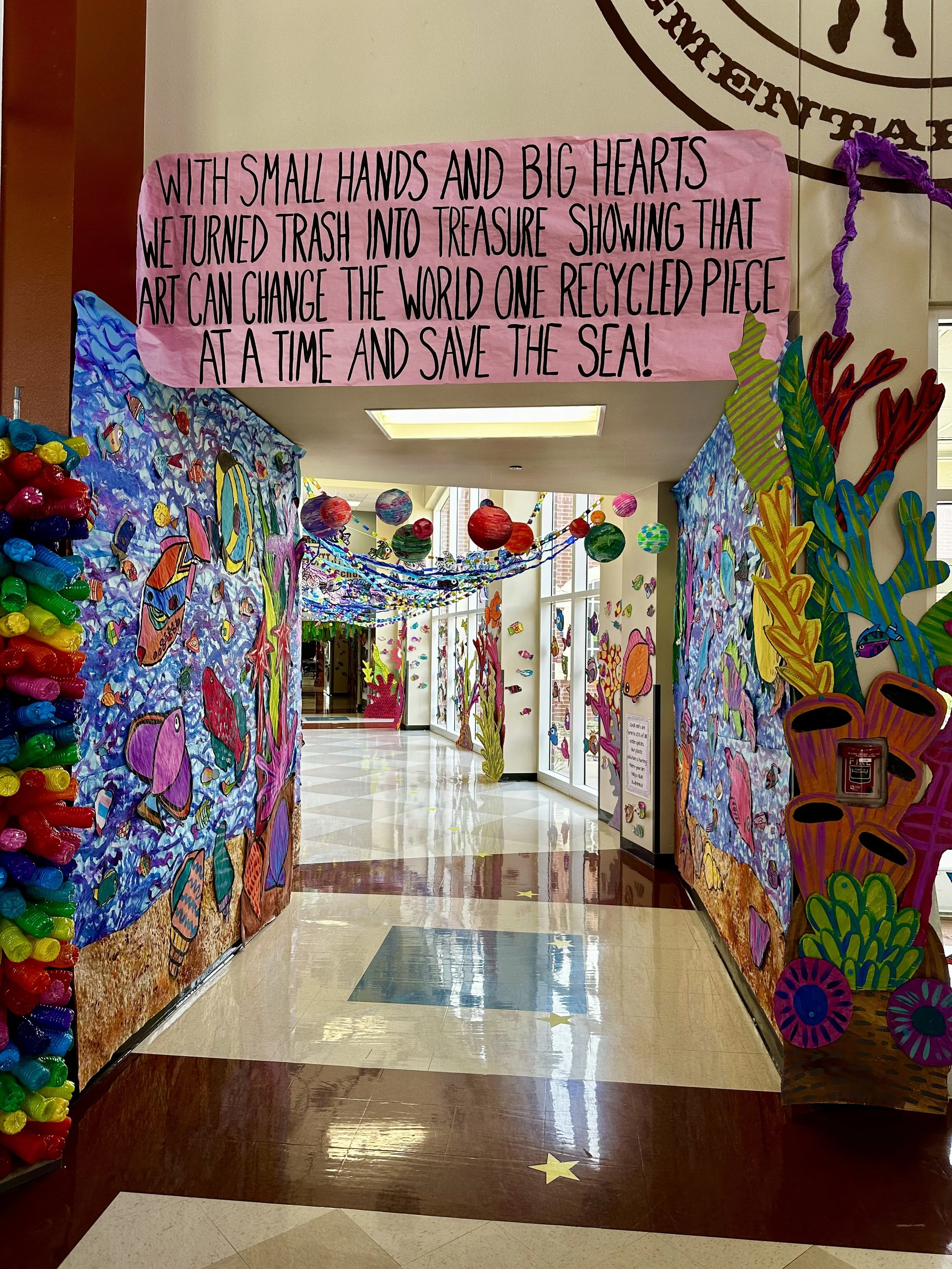 Colorful underwater-themed art display with a pink sign reading, 'With small hands and big hearts, we turned trash into treasure showing that art can change the world one recycled piece at a time and save the sea!'