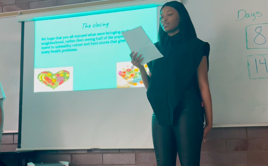 A young woman standing in front of a whiteboard giving a presentation, with slides projected behind her and some handwritten numbers on the whiteboard.