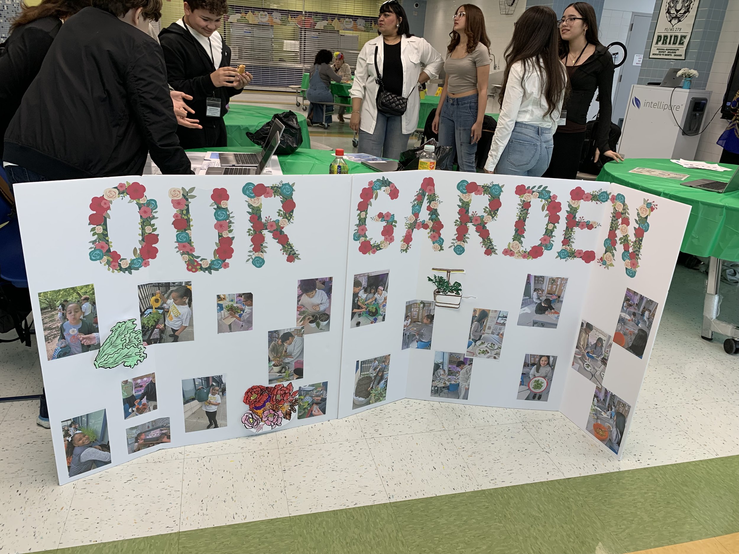 A white display board with the words 'Our Gardening' spelled out in large, colorful flower letters, featuring various photos of children and adults engaging in gardening activities, decorated with cartoon images of a watering can and flowers, set up at a community event inside a school or community center.
